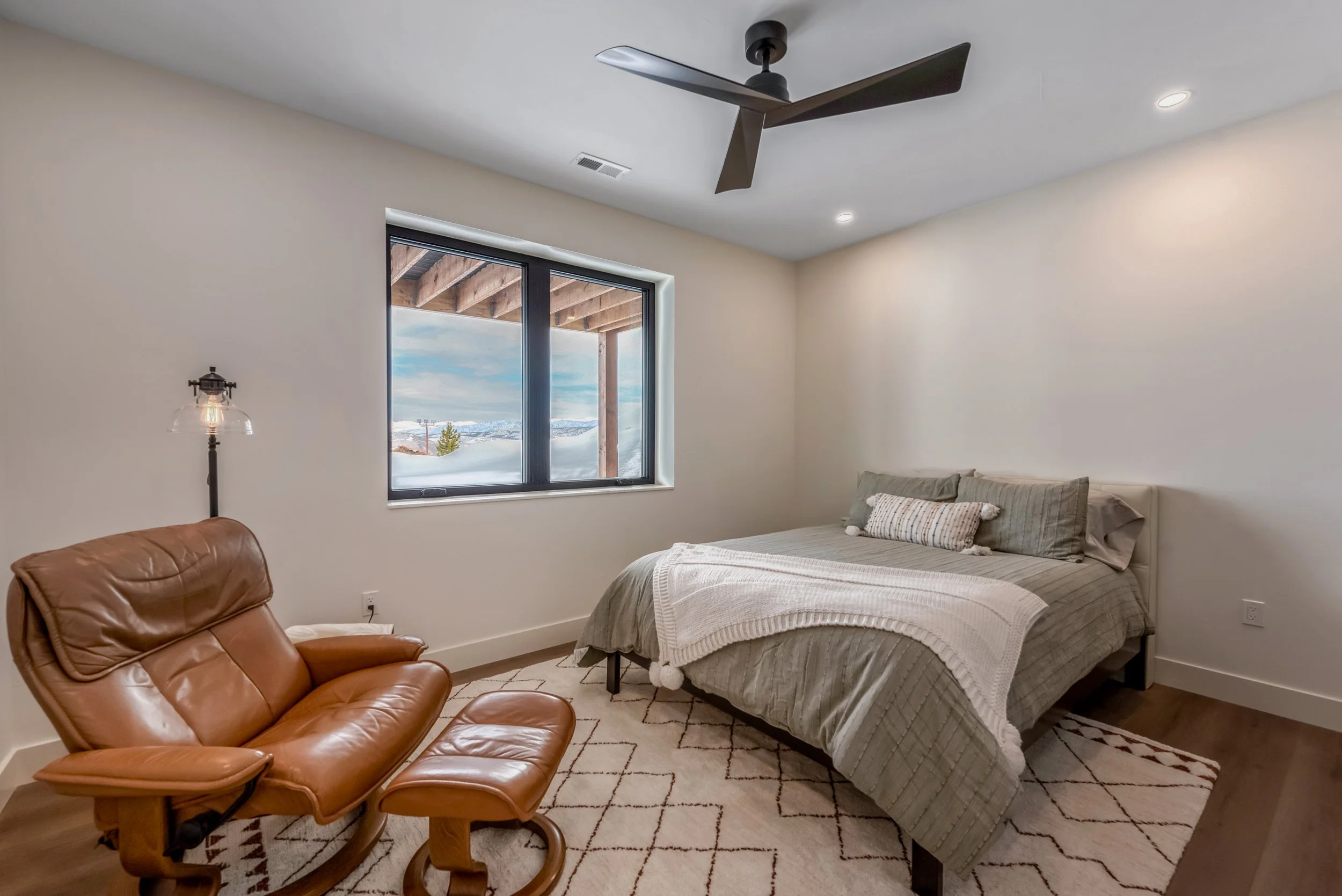 A bedroom with a large bed covered with beige and gray bedding, a brown leather lounge chair with a matching ottoman, a window showing a snowy landscape outside, and a ceiling fan.