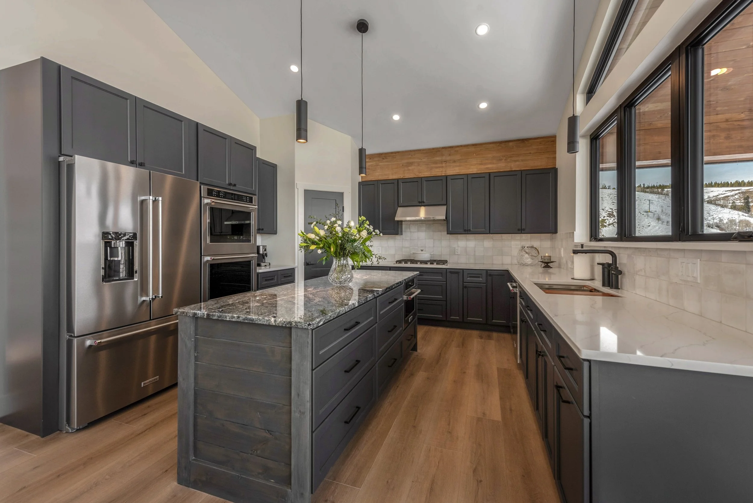 Modern kitchen with dark gray cabinets, stainless steel refrigerator, granite countertops, a kitchen island with a vase of flowers, large windows showing snow-covered landscape