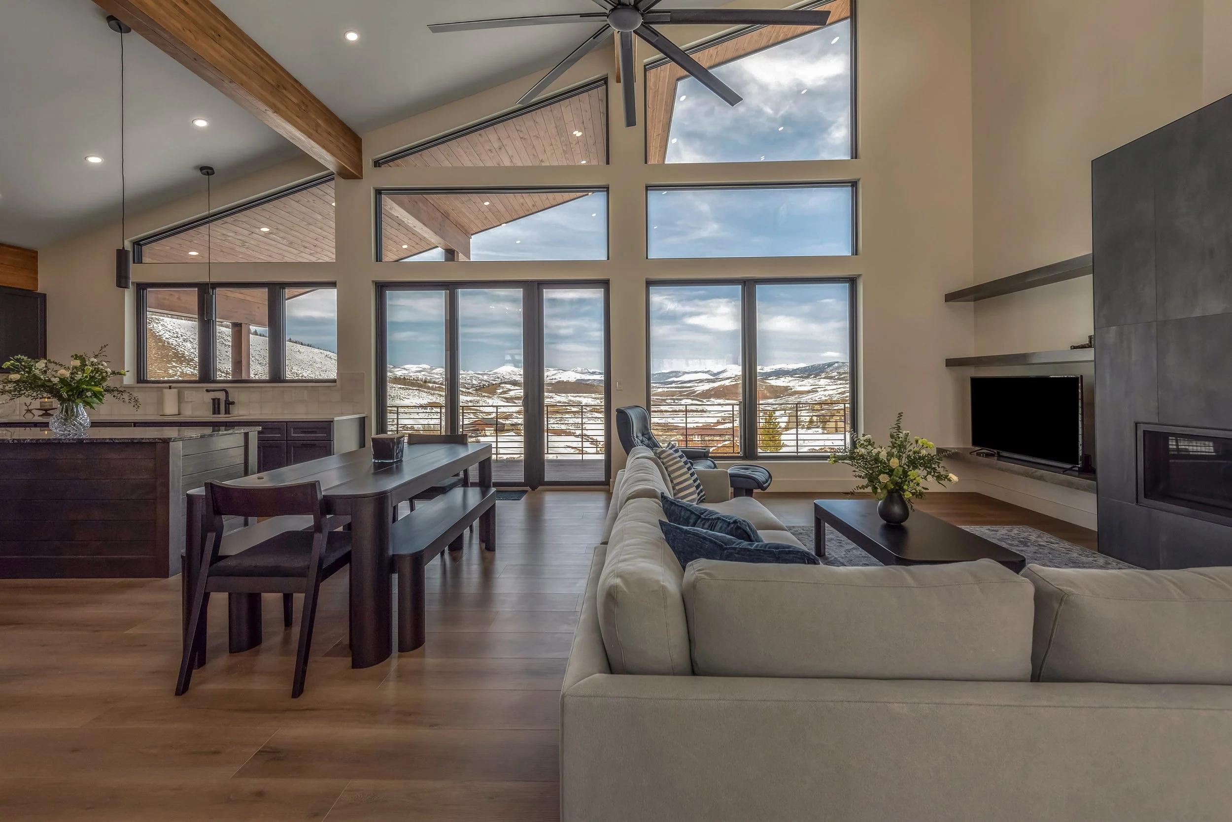 Living room with large floor-to-ceiling windows showcasing snowy mountain scenery, beige sofa with dark cushions, black dining table with matching chairs, modern fireplace, TV, ceiling fan, and wooden beams on the ceiling.