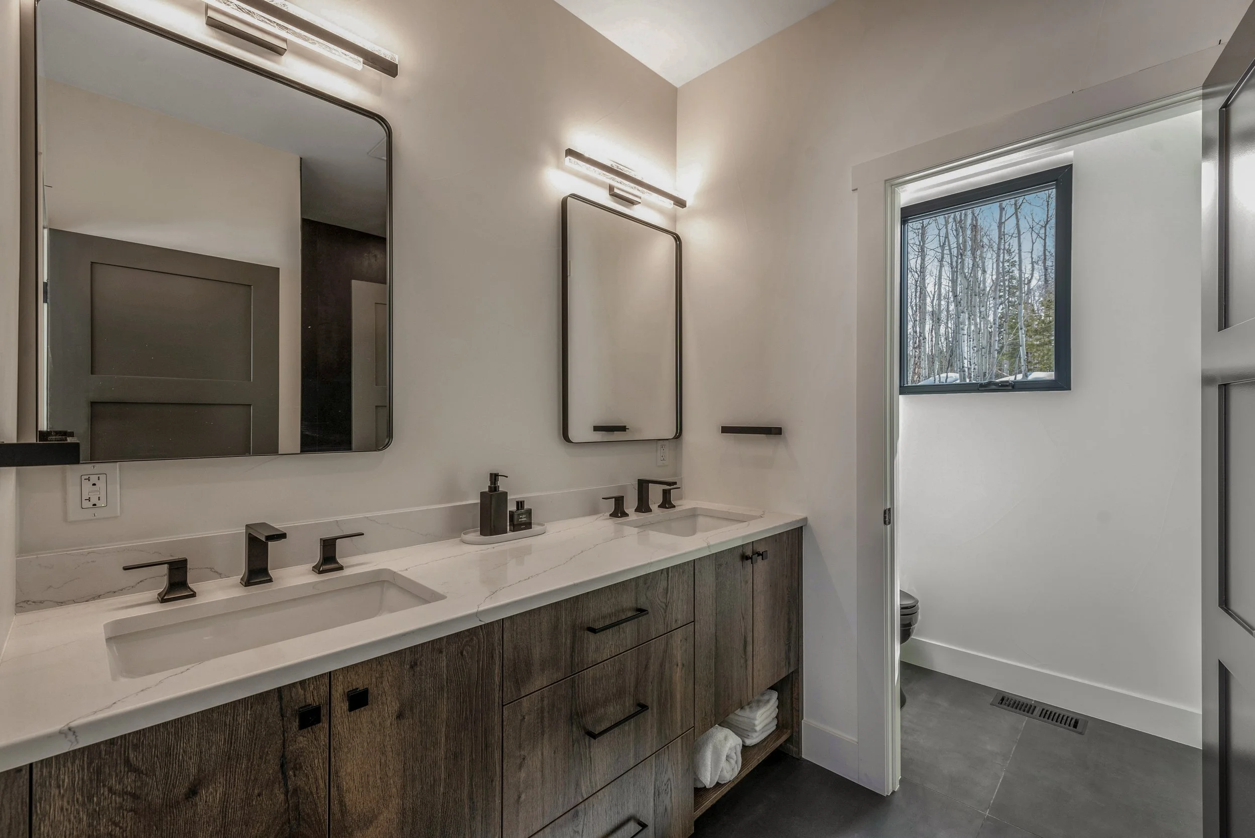 Modern bathroom with dual sinks and wooden vanity, white marble countertop, black fixtures, large mirror, and a separate toilet area with window and natural light.