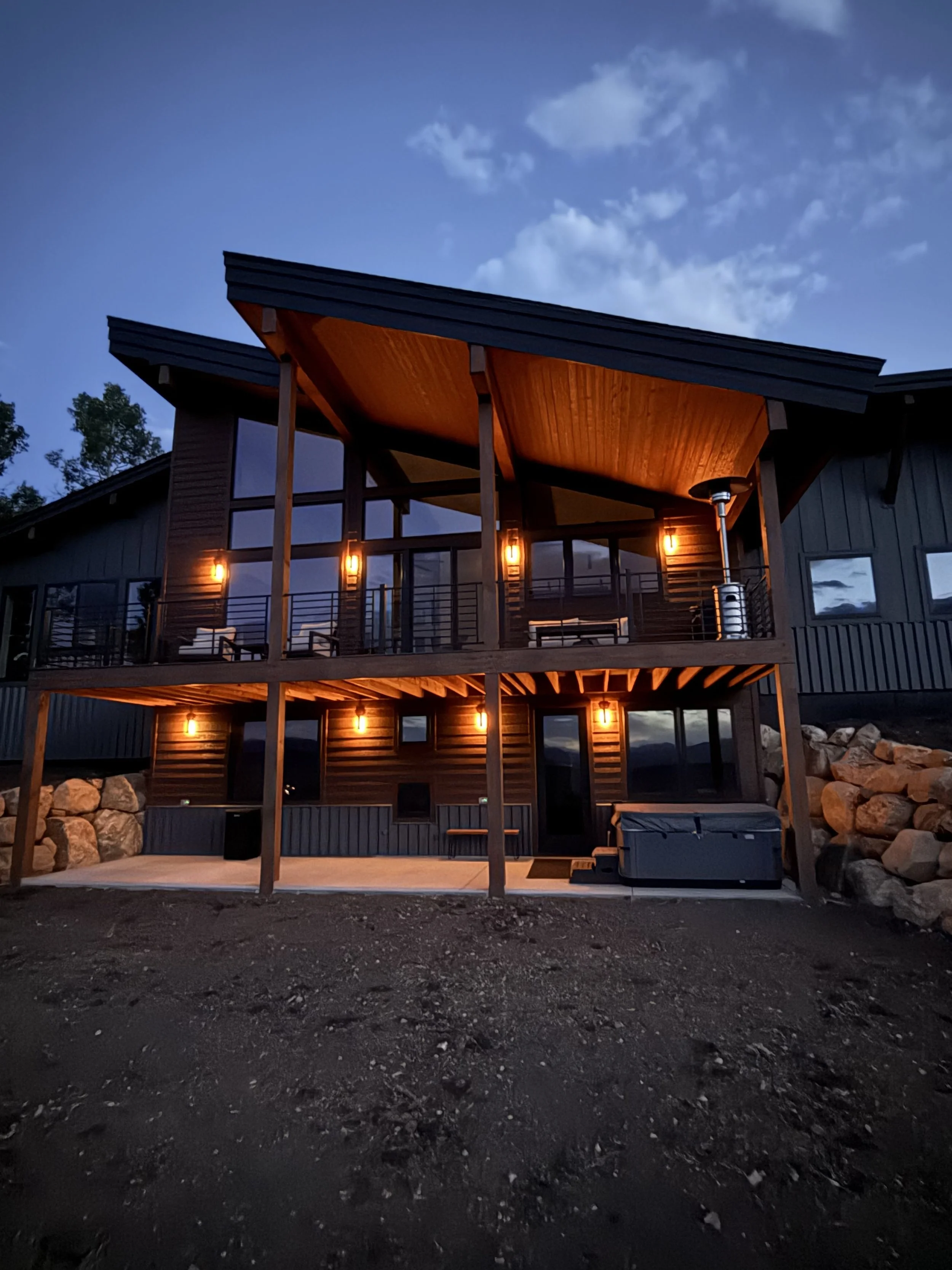 A modern house with a two-story porch, illuminated with warm outdoor lights, in a natural setting during evening with a partly cloudy sky.