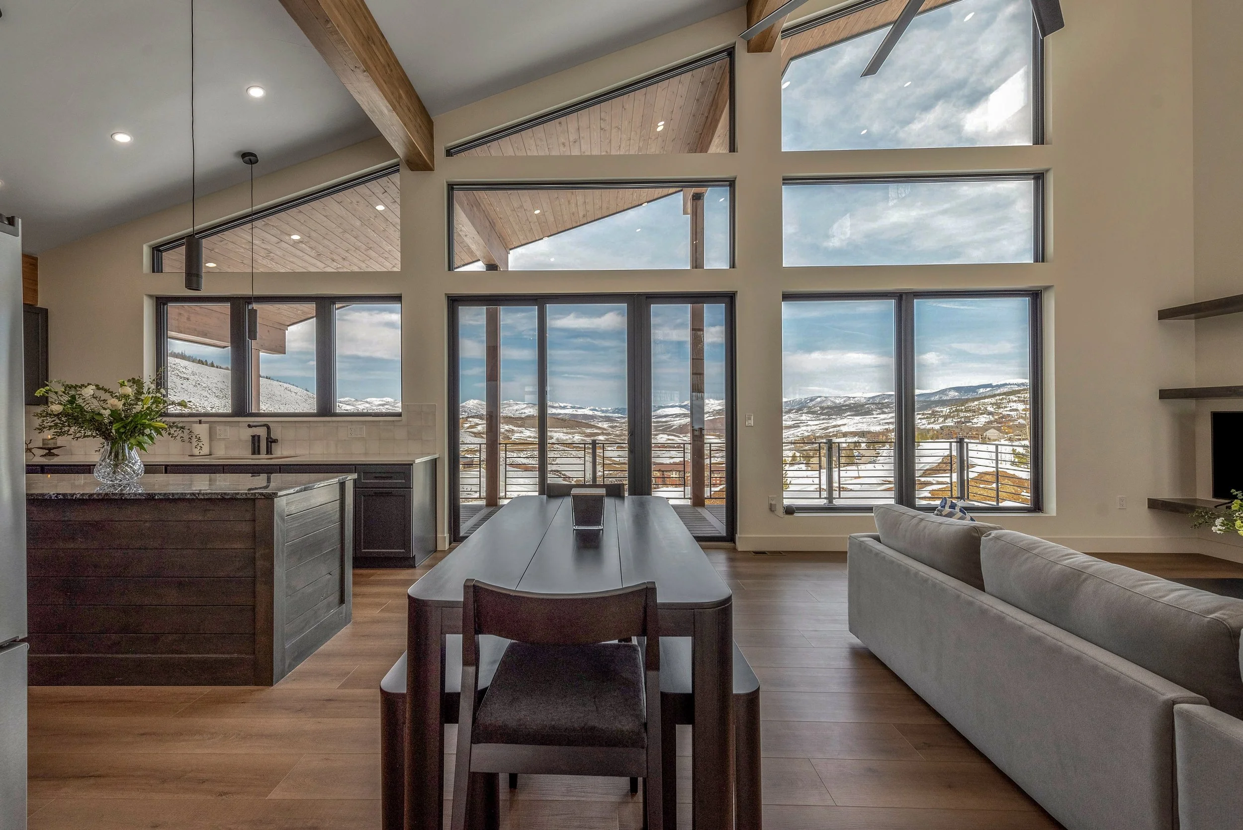 Living room with large windows and sliding glass door overlooking snow-covered mountains, modern decor, dining table, and gray sofa.
