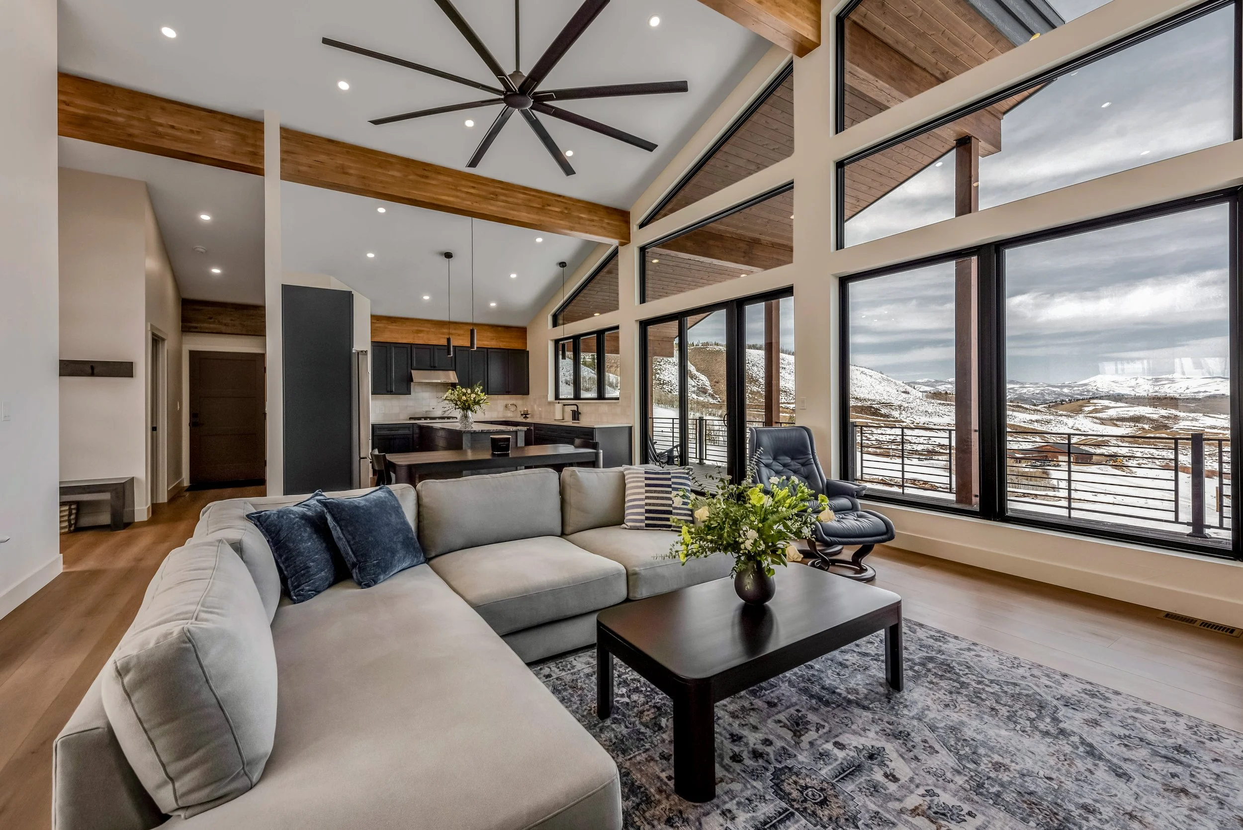 Living room with large windows showing snowy mountain landscape, beige sectional sofa with dark pillows, black armchair, dark wood coffee table with flower arrangement, open kitchen with black cabinets, and wooden ceiling beams.