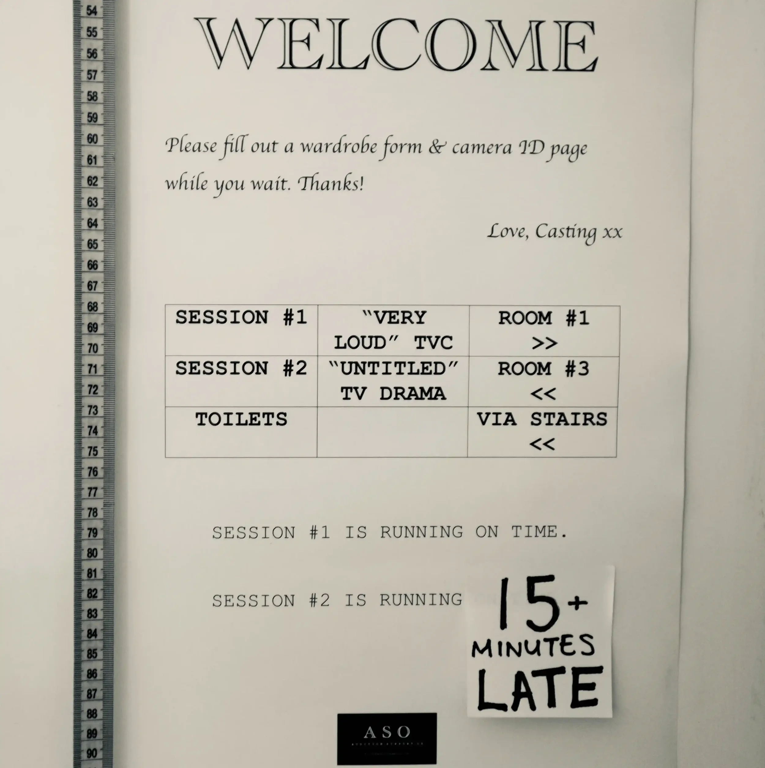 A "Welcome" sign in a casting office hangs on the wall accompanied by a soft measuring tape. The sign suggests there is a casting for a loud TV commercial & an untitled TV drama series taking place at the same time.