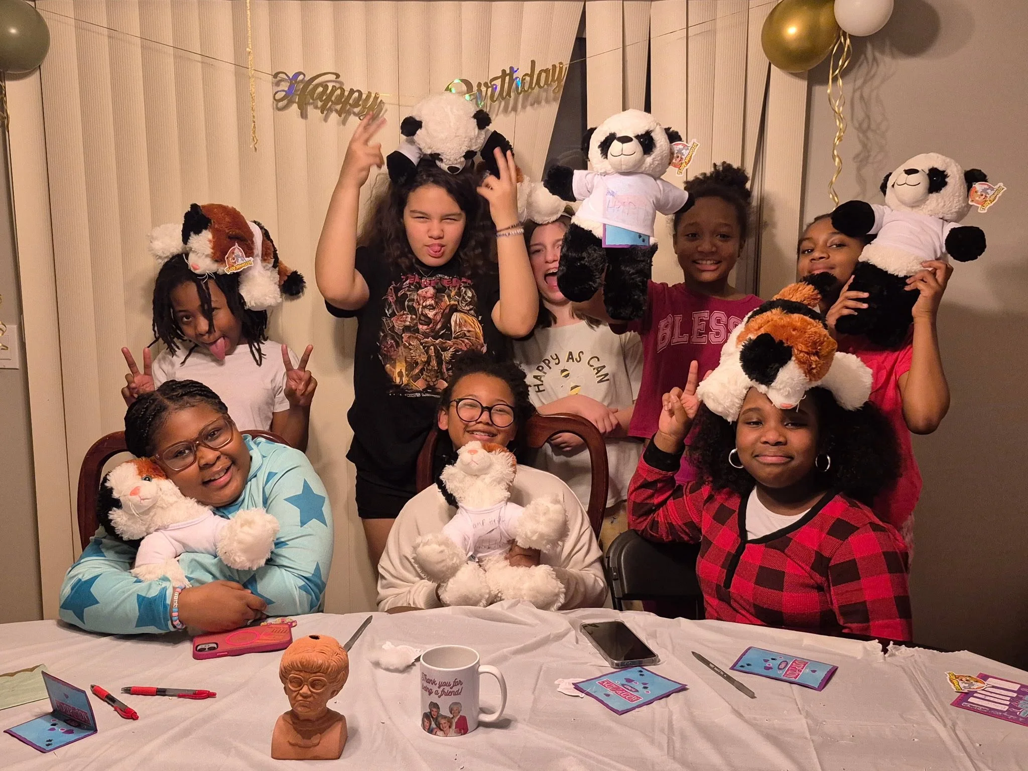A group of children celebrating a birthday, holding stuffed panda and dog toys, sitting and standing around a table with party supplies, smiling, making silly faces, and showing peace signs.