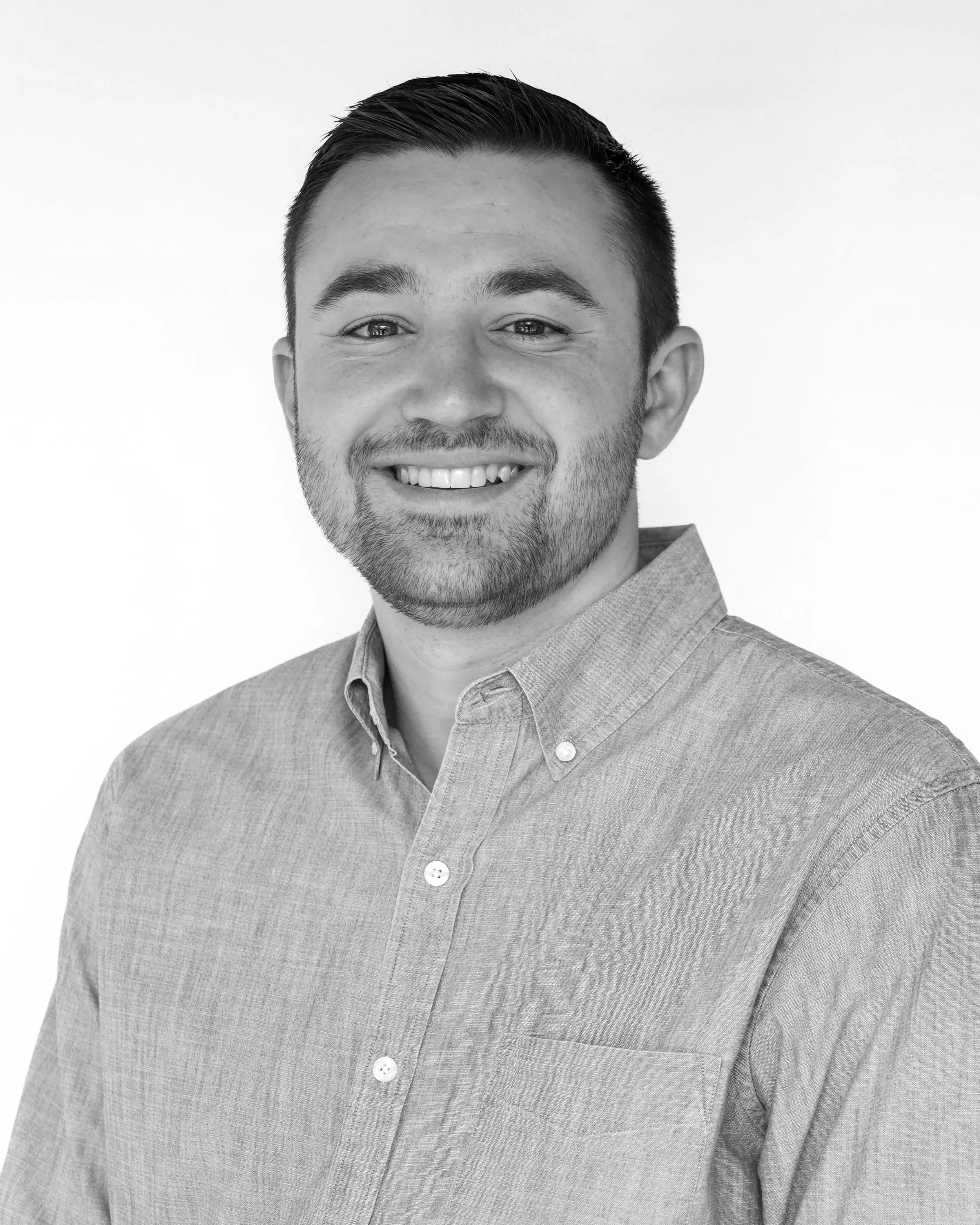 A smiling man with short dark hair, a beard, and a mustache, wearing a button-up shirt, posing against a plain background.