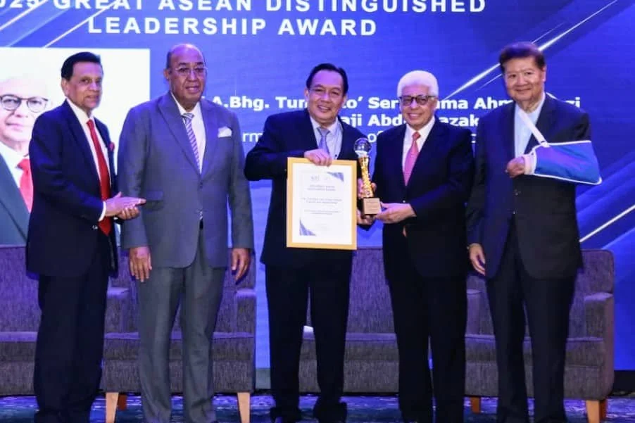 Five men in formal suits on stage at an award ceremony, with one man holding a framed certificate and a trophy, and the background displaying text about the ASEAN Leadership Award.