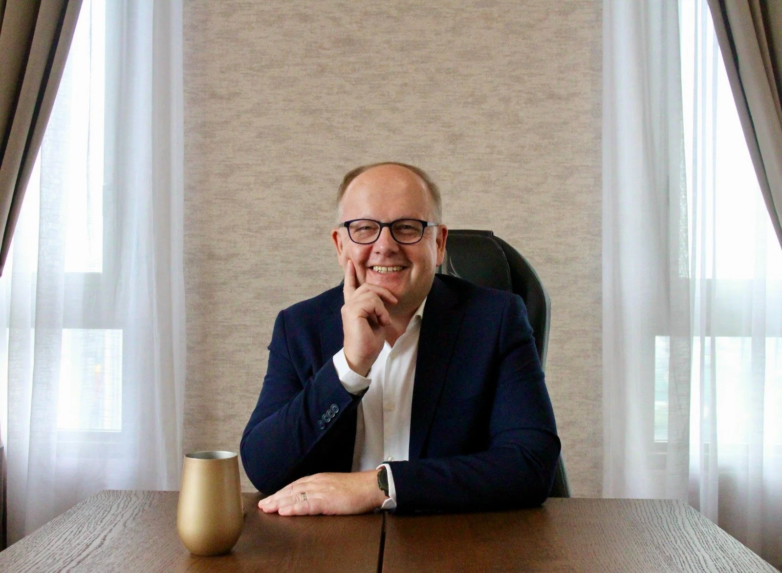A smiling man with glasses wearing a dark blazer and white shirt, sitting at a wooden table in an office, with a gold mug in front of him and curtains in the background.