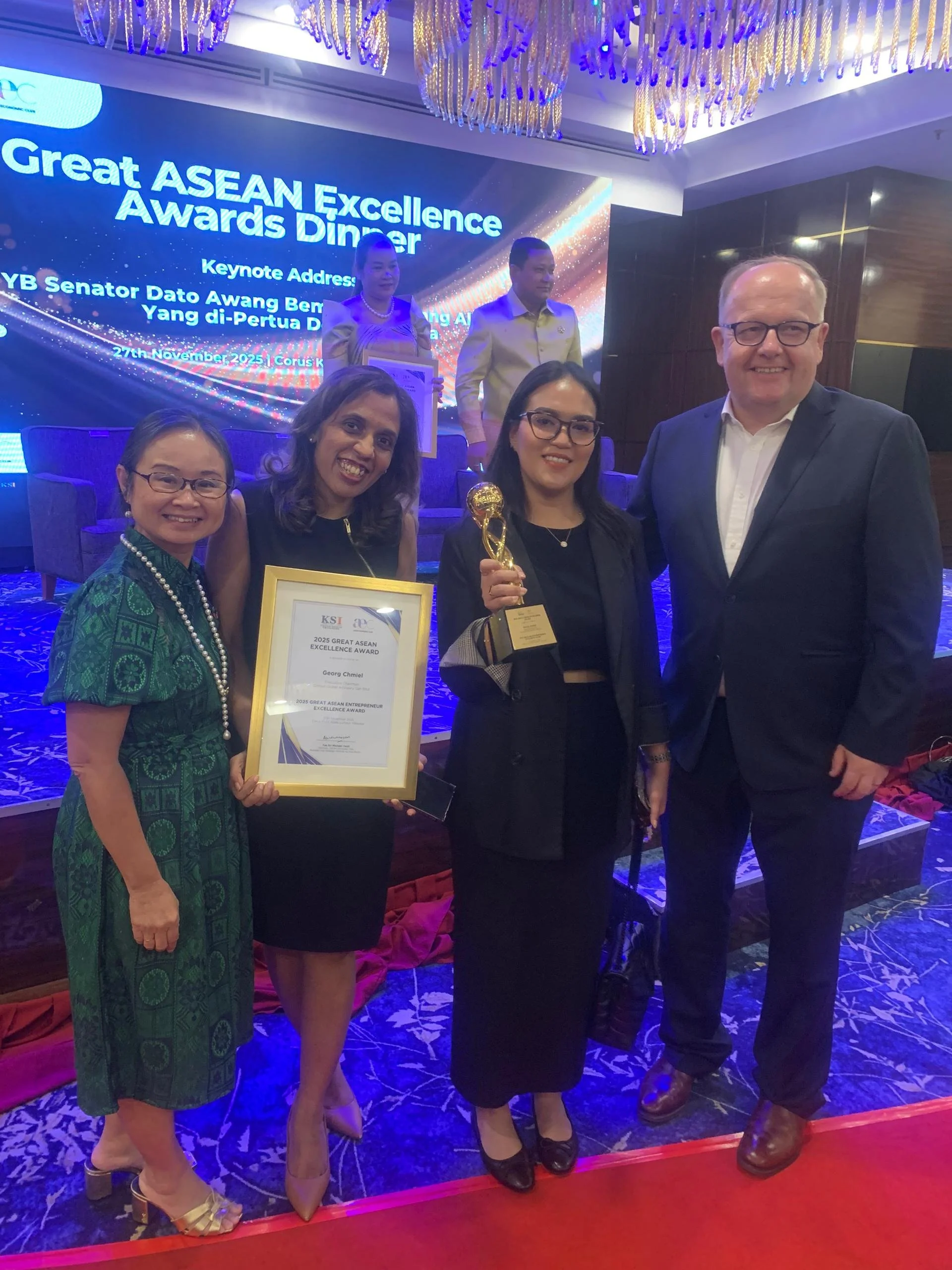 Group of five people standing together on stage at an awards dinner, with a woman holding a framed certificate and another woman holding a trophy. Behind them is a large digital screen displaying event information and a couple of people standing on a