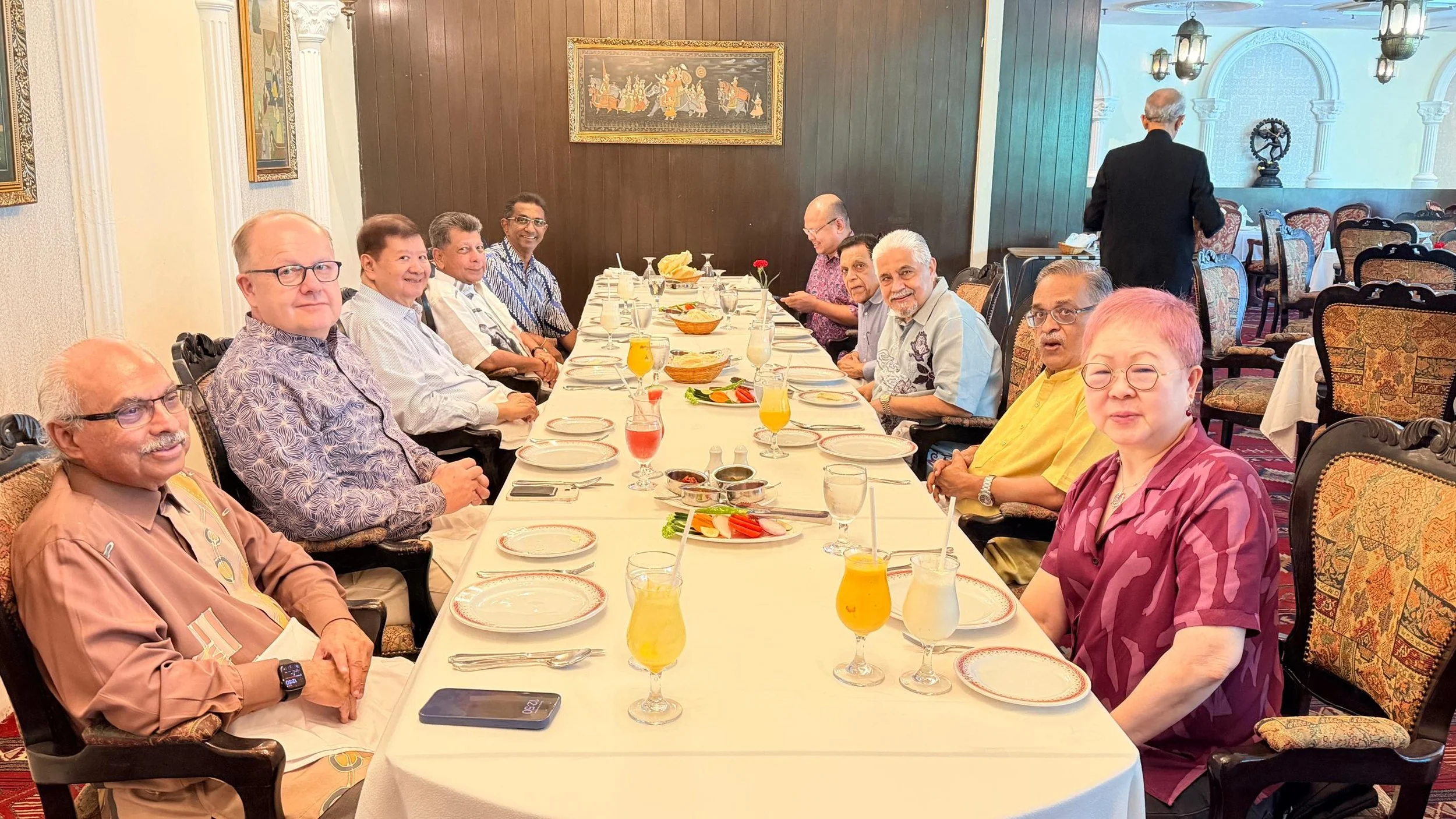 A group of people seated around a long dining table in a restaurant, with plates, glasses of drinks, and food on the table, preparing for a meal.