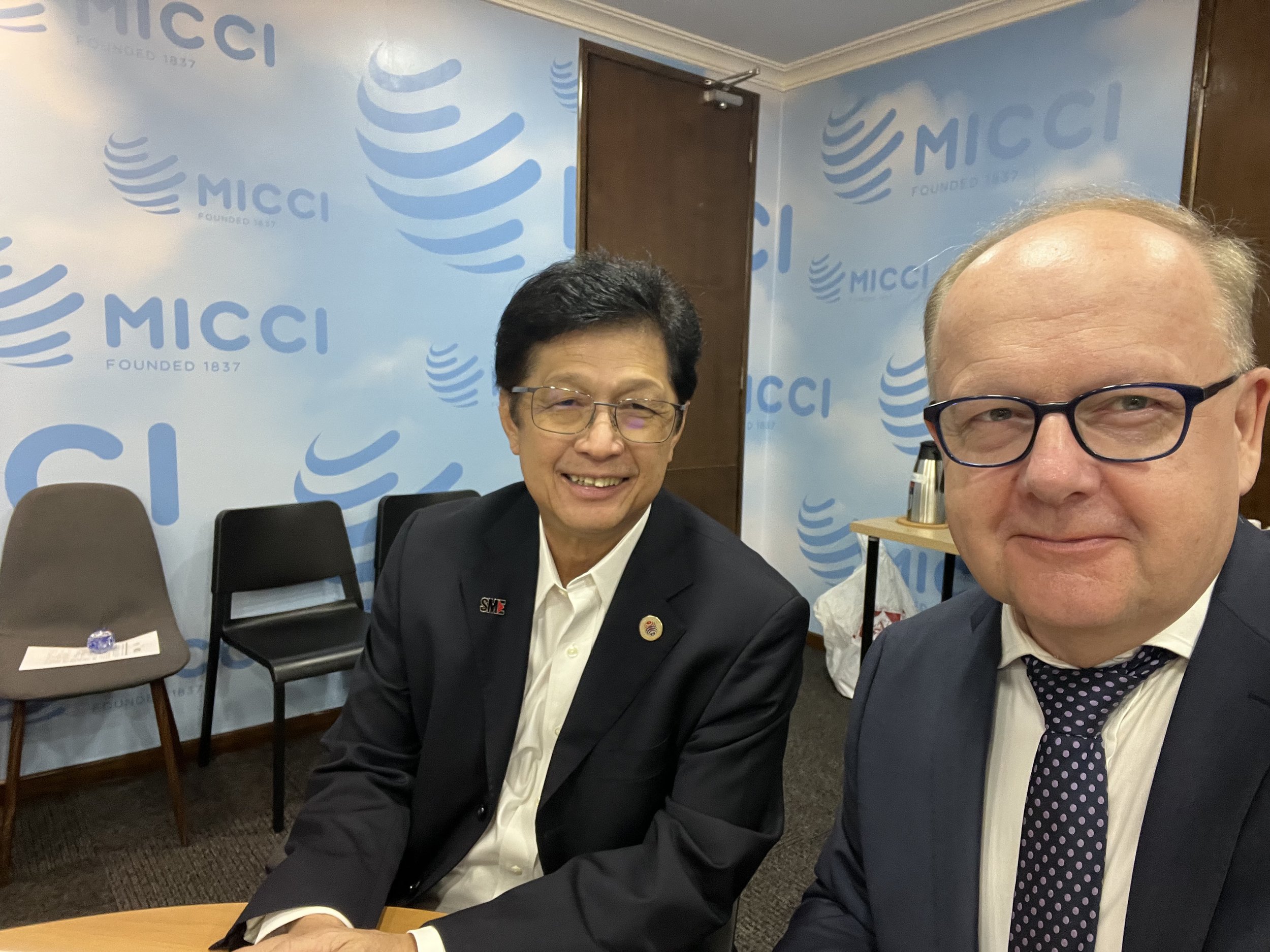 Two men in business suits sitting at a table, smiling for a photo, with a blue wall in the background that has the MICCI logo and the words 'Founded 1837'.