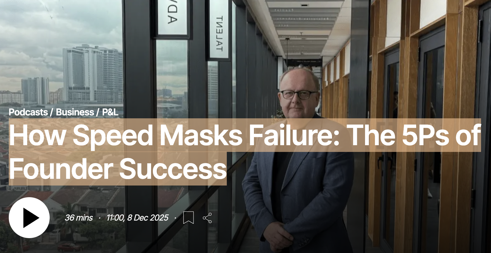 A man with glasses in a suit standing in a modern office corridor with large windows, cityscape view, and wooden panel walls.