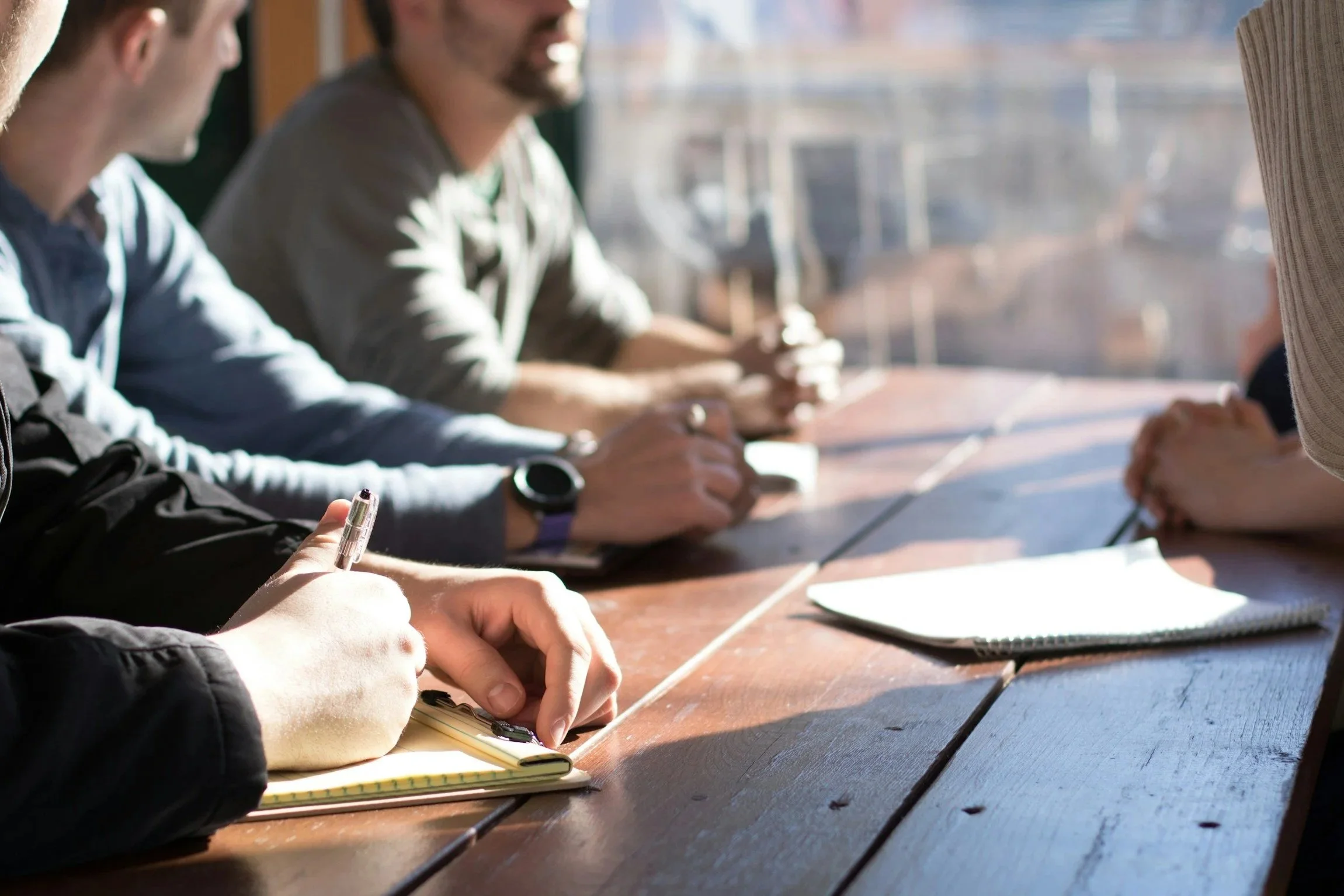People sitting at a wooden table, engaged in a discussion or meeting, with notebooks and pens, in a bright room with natural sunlight.