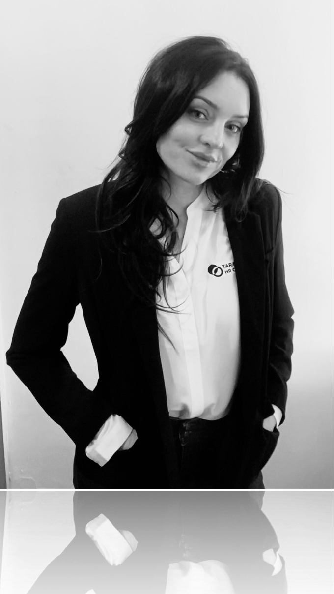 Tara Byrnes - A woman with long dark hair wearing a black blazer and white shirt with a company logo, standing with hands in pockets against a plain background.