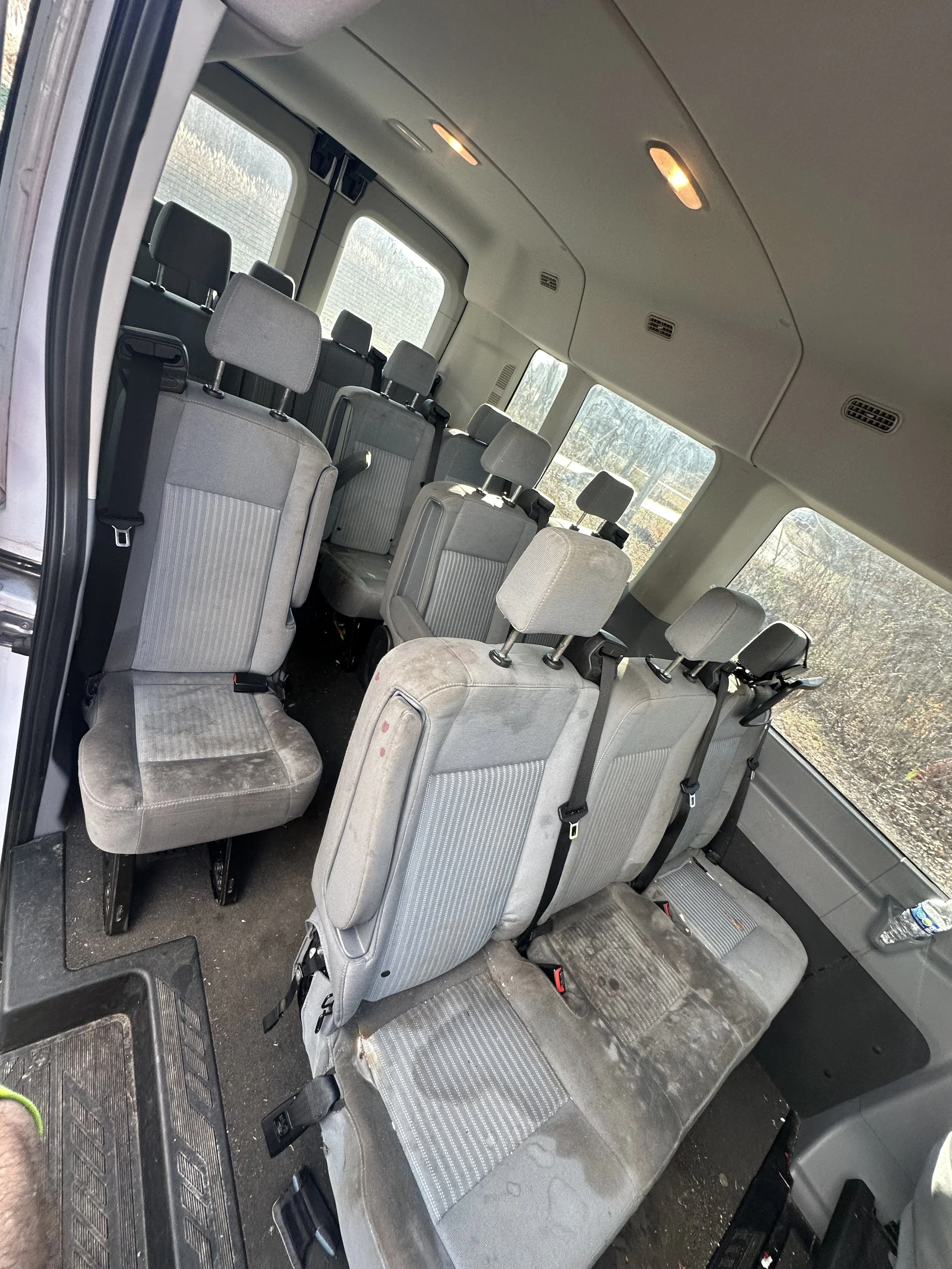 Interior of a passenger van with multiple gray fabric seats, some stained and dirty, and a water bottle on the side panel.