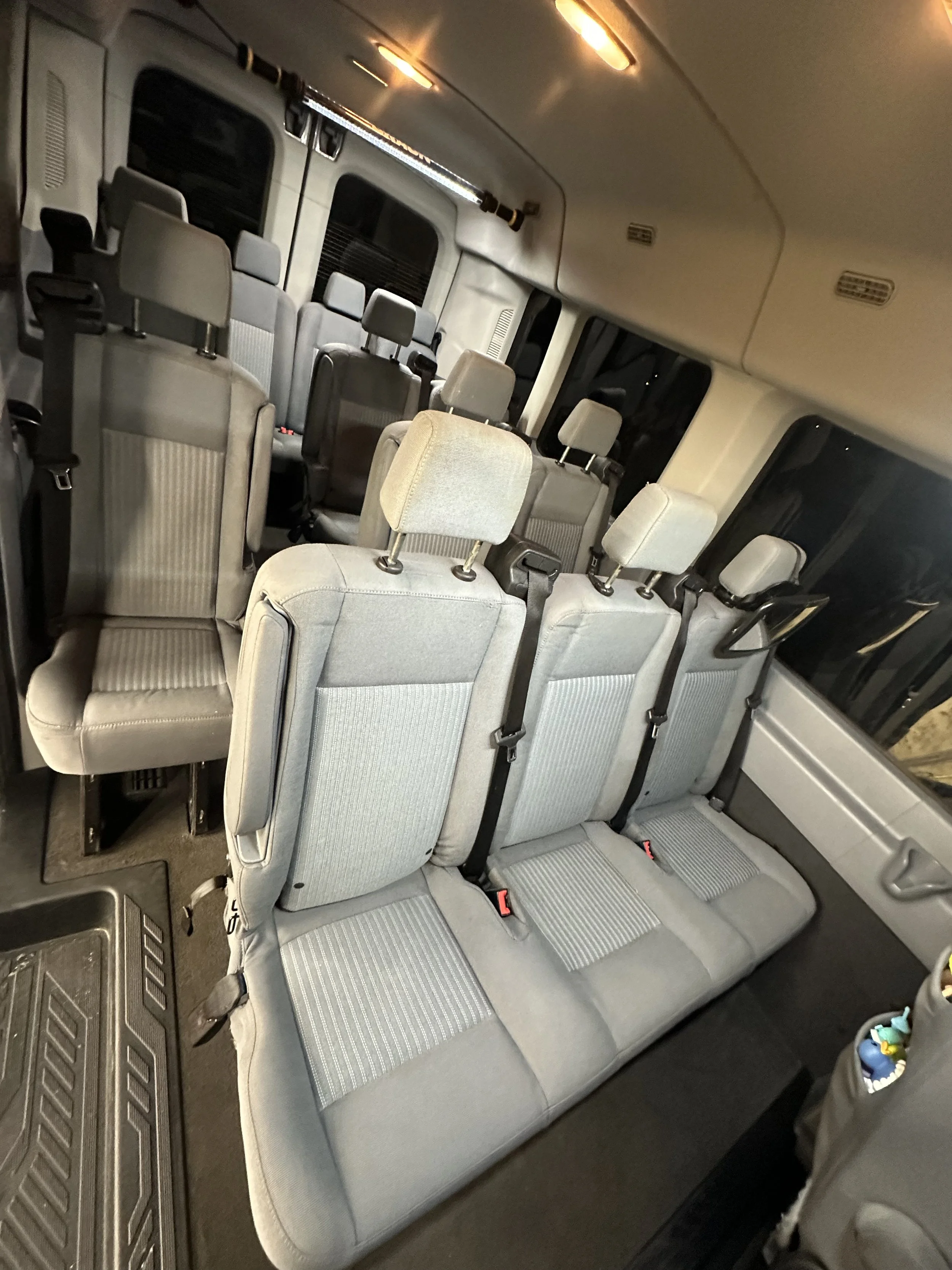 Interior of a vehicle showing multiple rows of gray fabric seats with seat belts, window, and overhead lights.