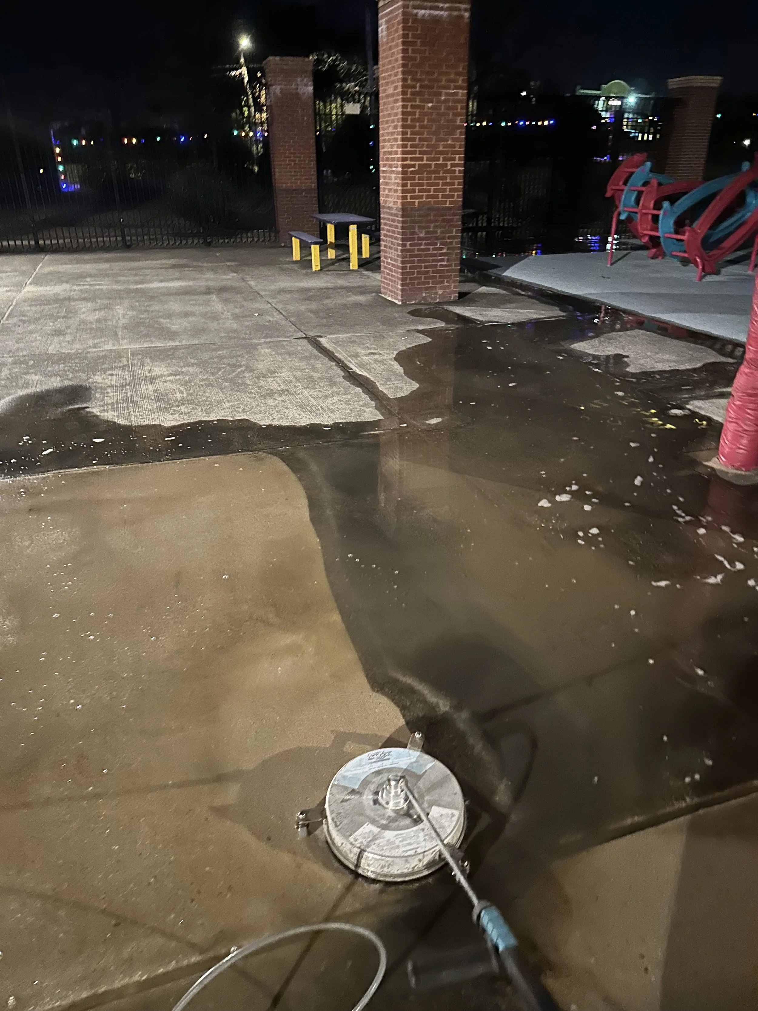 Nighttime scene of a wet sidewalk with a floor buffer or polisher in the foreground, near a brick pillar and a metal fence, with colorful lights in the background.