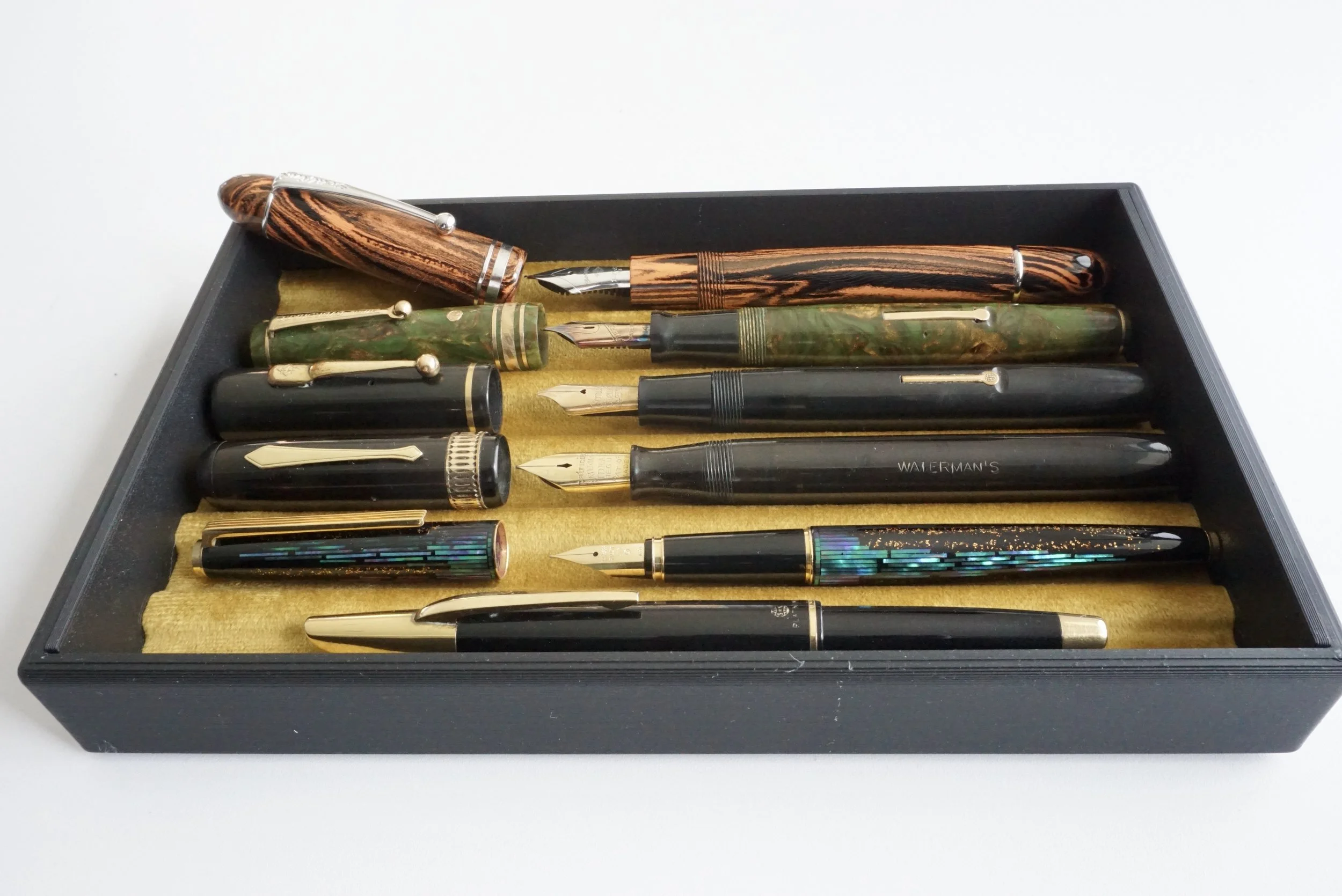 A black box containing a collection of vintage fountain pens with various colored and patterned bodies, some with gold accents and nibs, arranged on a golden lining.