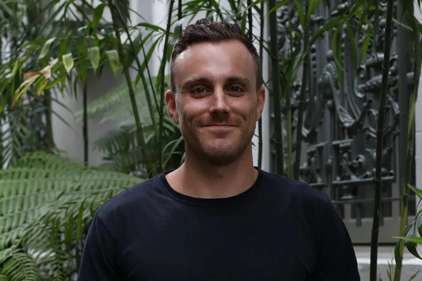 A man smiling in front of tropical plants and a metal gate.