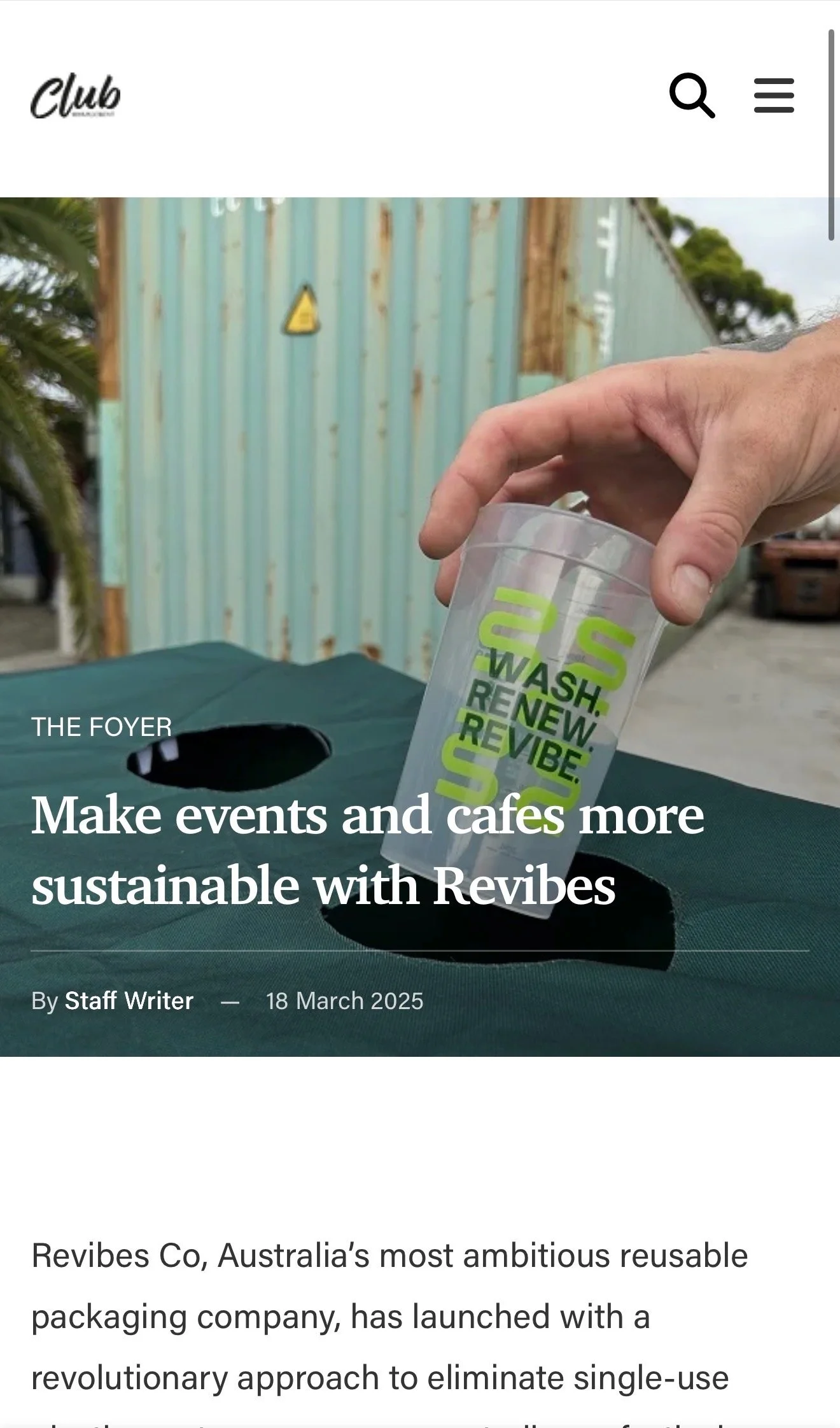 A clear plastic reusable cup with green and black text that reads '2 WASH. RENEW. REVIVE'. The cup is held by a person's hand over a dark green table with two large circular holes. In the background, there is a light blue corrugated metal wall with rust spots and a yellow triangular warning sign, outdoors with some trees and a parked vehicle visible.