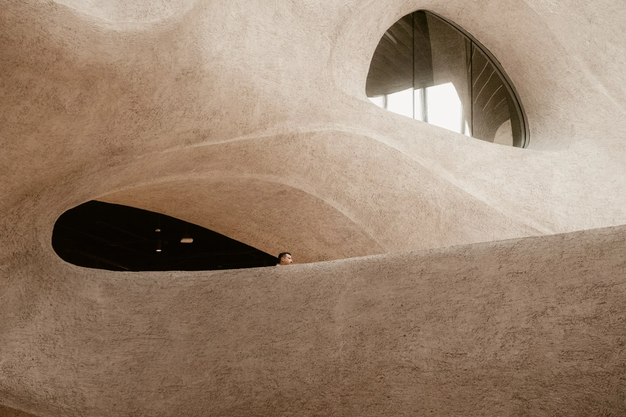 Interior of a sculptural beige concrete building with curved walls, arched windows, and a person visible through an opening