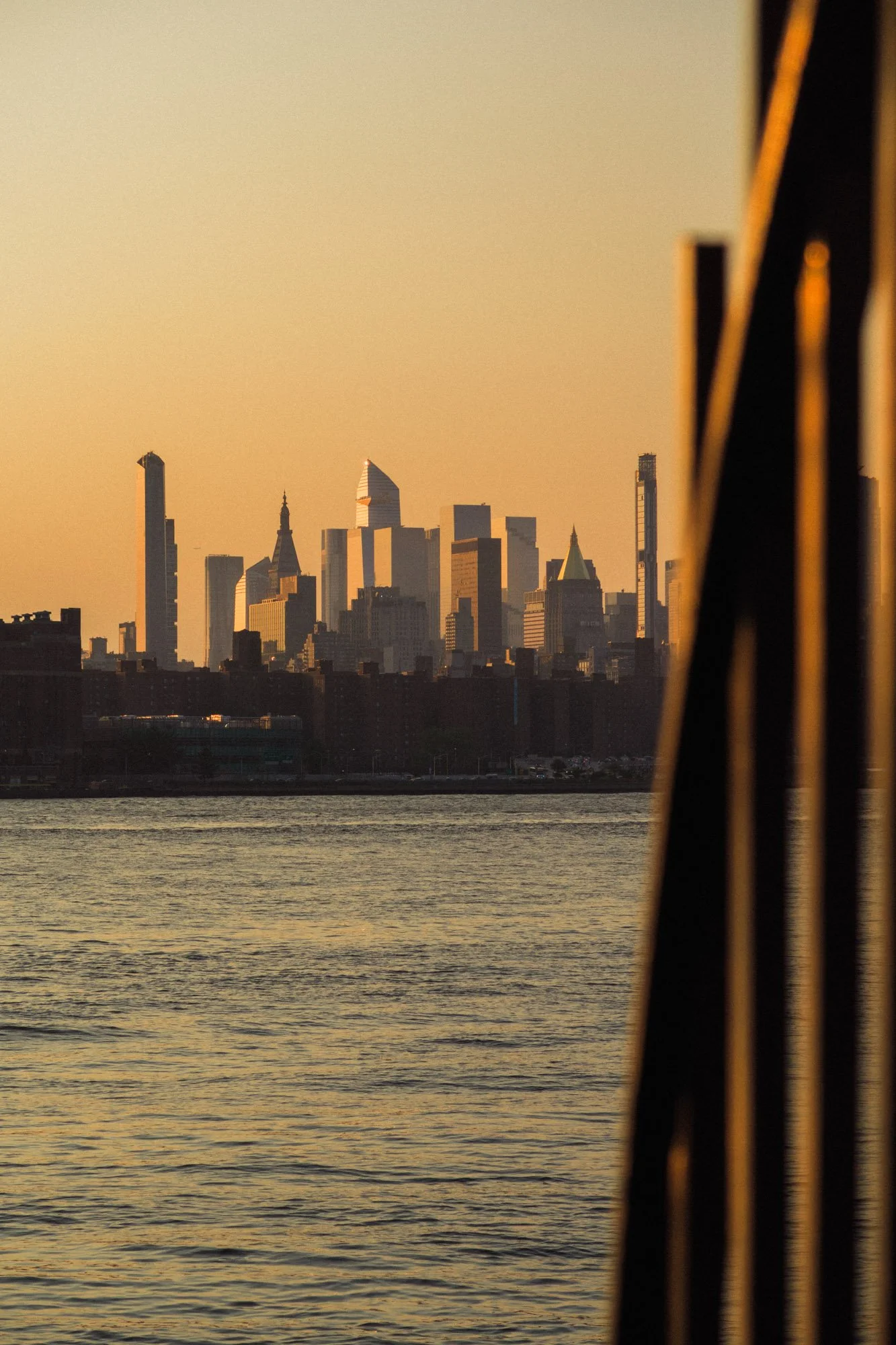 New York City skyline at sunset viewed across the Hudson River, framed by the Brooklyn Bridge cables.