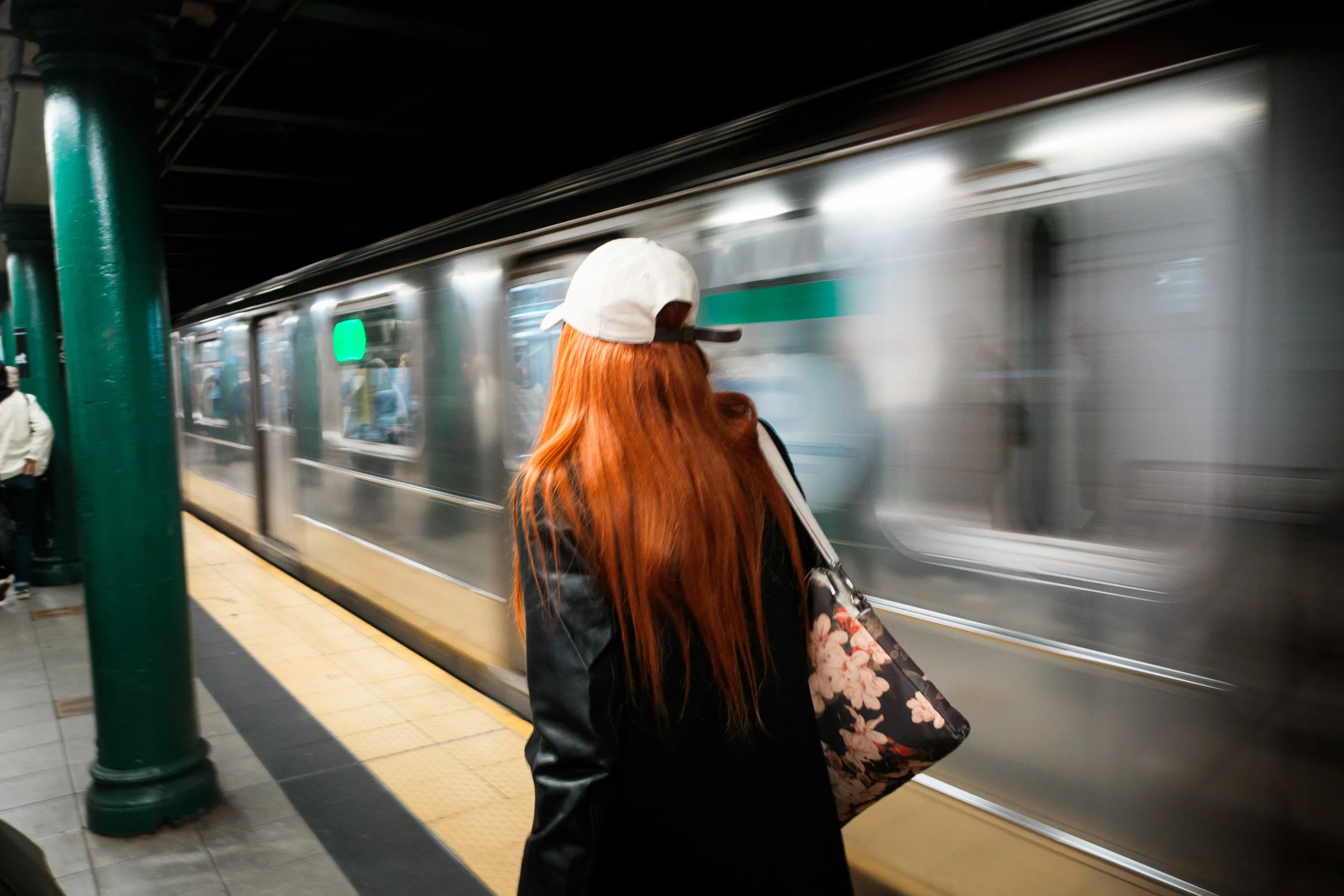 Person with long red hair waiting on a subway platform as a train speeds past