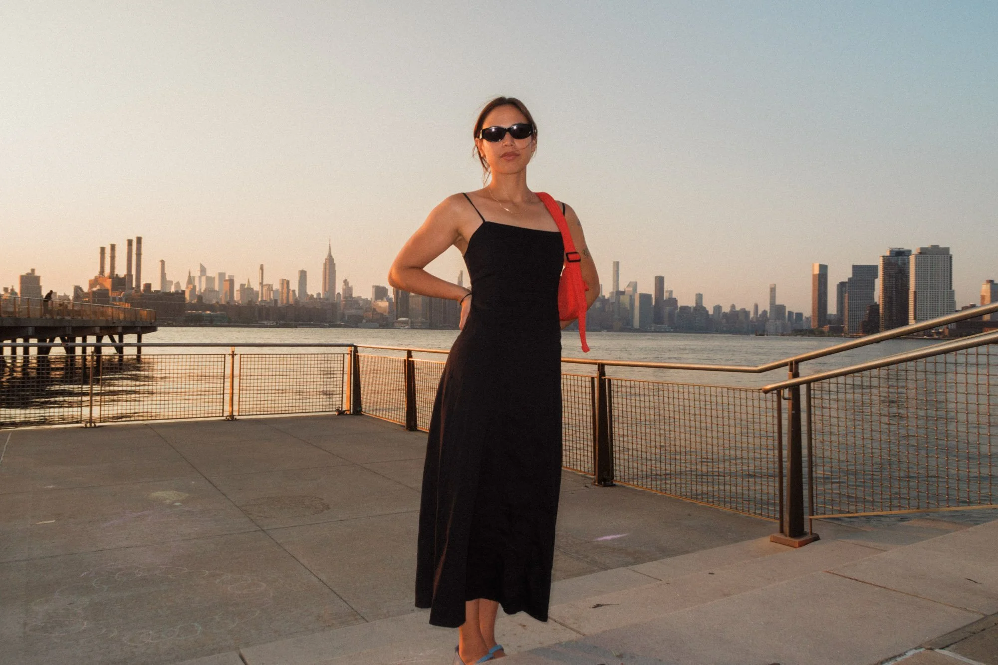 Woman in a black dress and sunglasses posing on a waterfront promenade with the Manhattan skyline at sunset