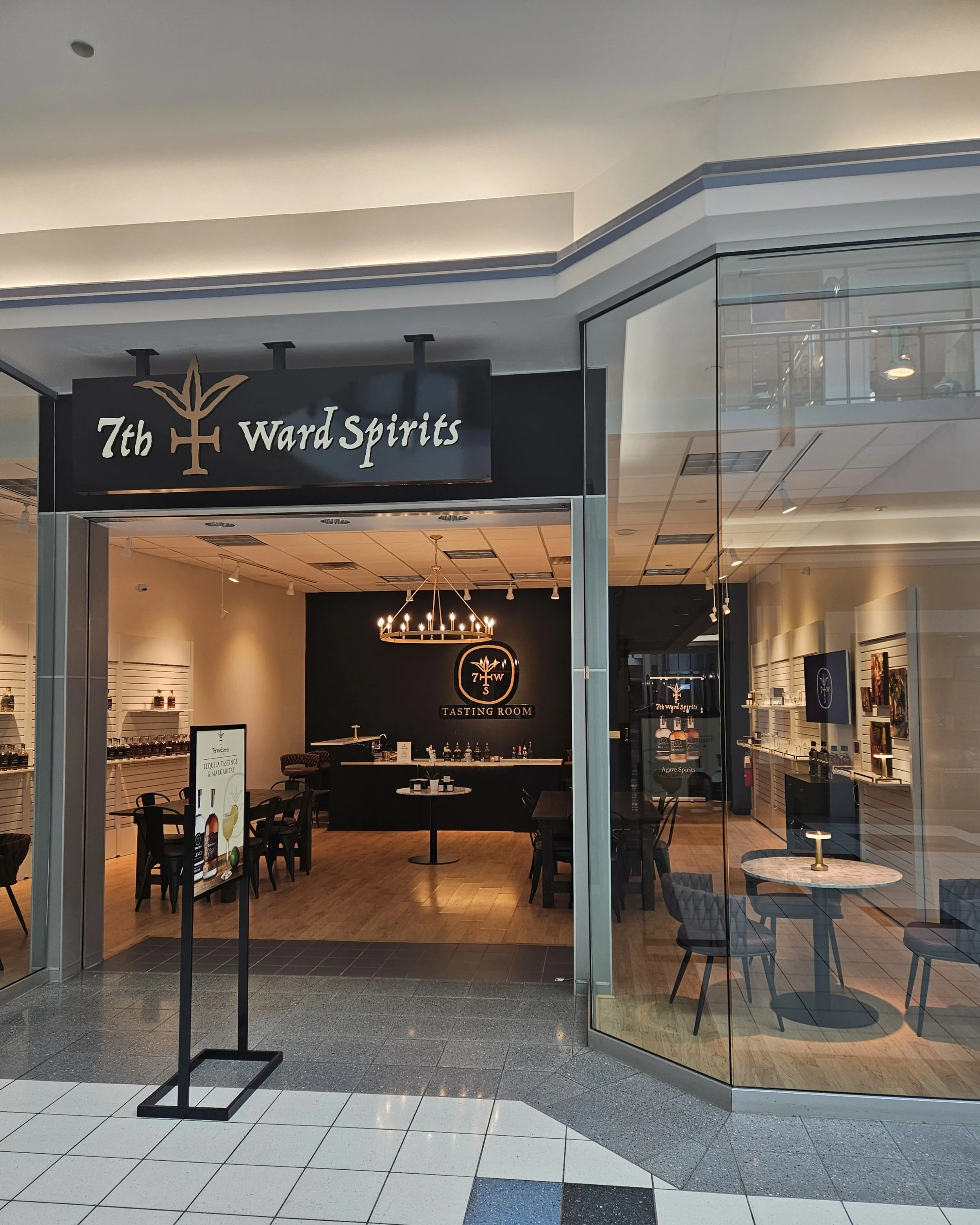 View of the entrance to 7th Ward Spirits tasting room inside a shopping mall, with black sign featuring brown and white lettering, glass walls with reflections, and a cozy interior with tables, chairs, and decorative lighting.