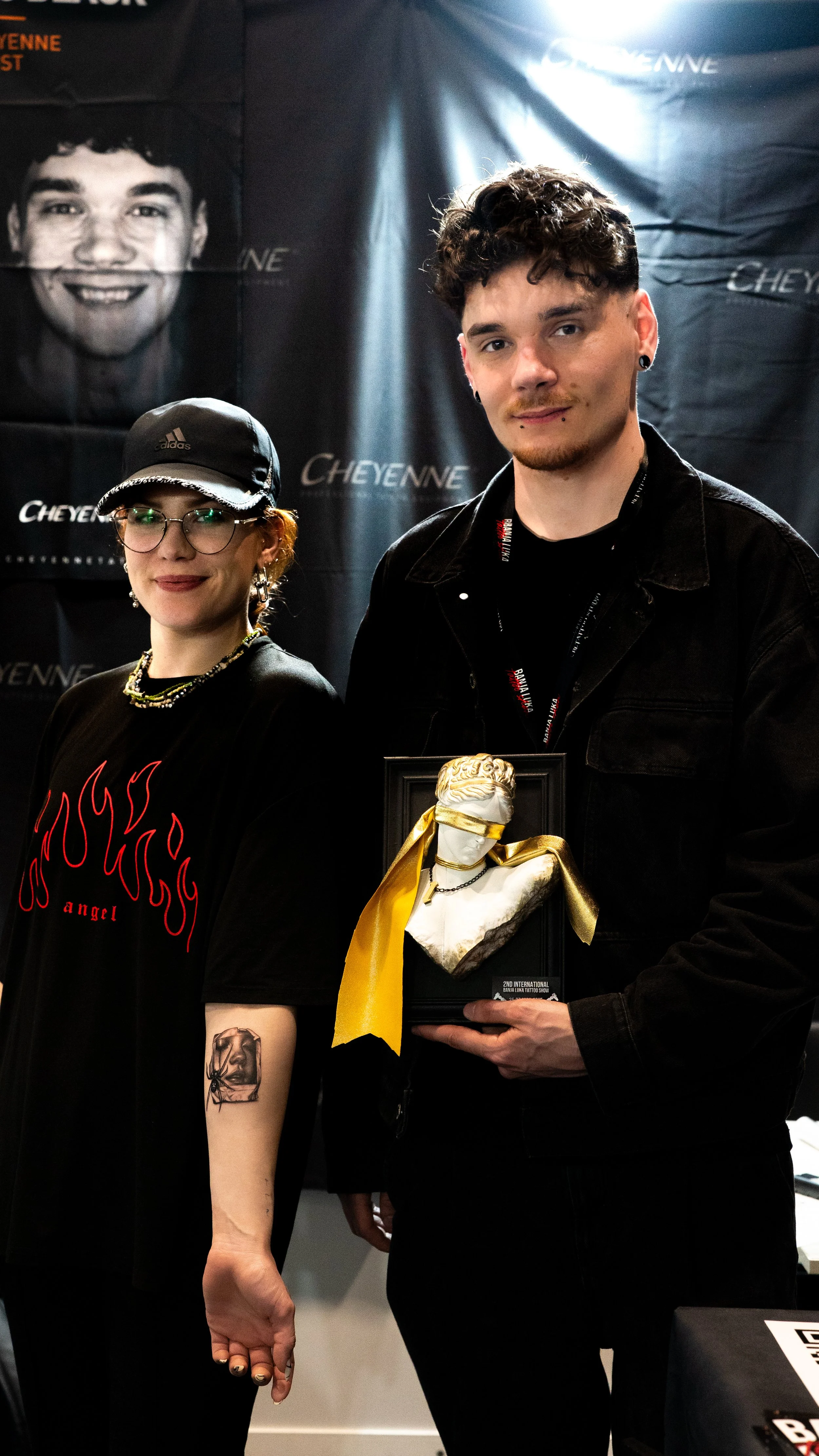 Zwei Personen stehen vor einem Cheyenne-Banner und halten eine Skulptur einer Frau mit verbundenen Augen und einem goldenen Band in den Händen. Die Person auf der linken Seite trägt eine Brille, eine schwarze Mütze und ein schwarzes T-Shirt mit roten Flammenmotiven sowie ein Tattoo am Unterarm. Die Person auf der rechten Seite hat kurzes, lockiges Haar, mehrere Ohrringe und ist ganz in Schwarz gekleidet; um den Hals trägt sie ein Schlüsselband.