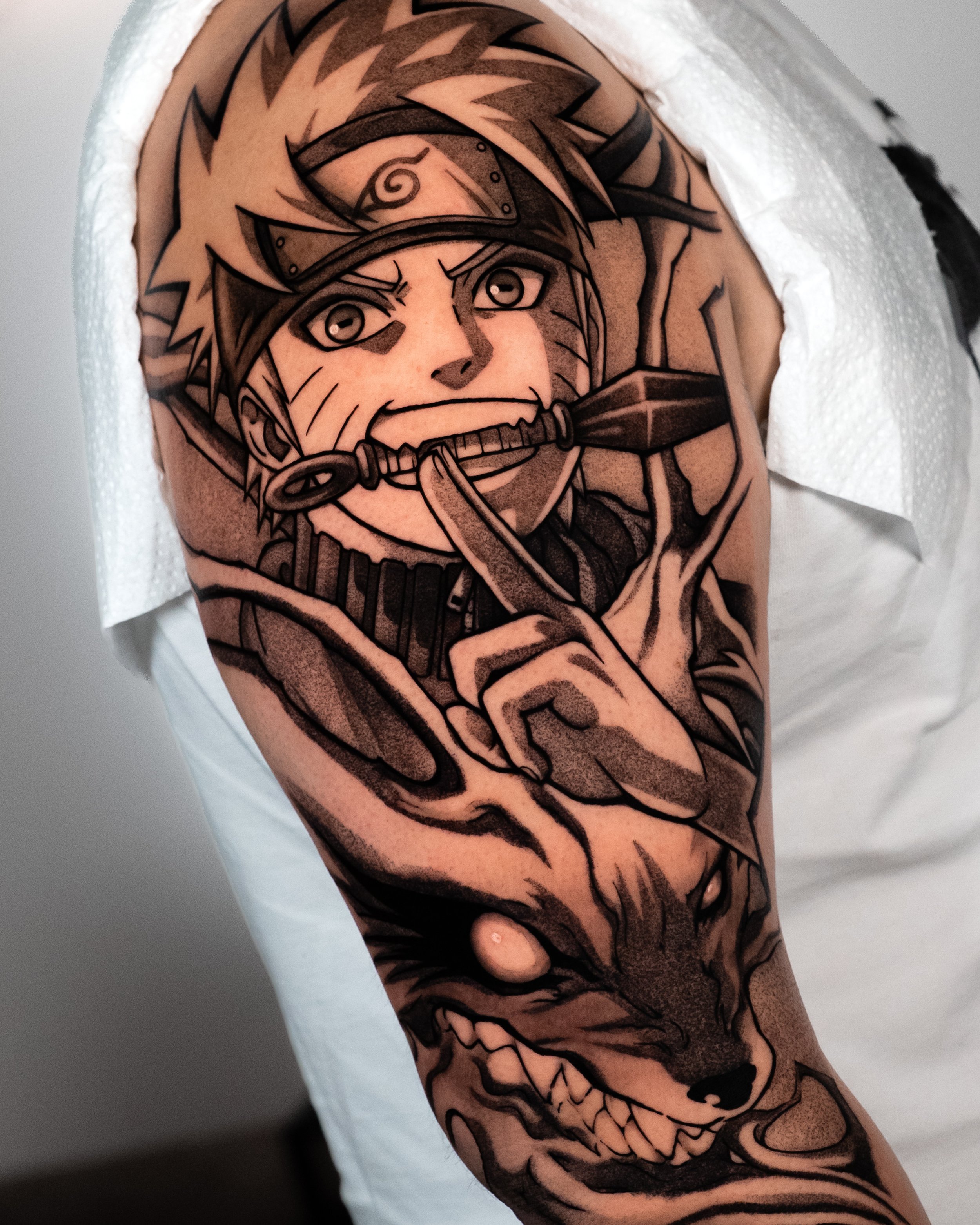 Tattoo of a fierce anime-style character with spiky hair, headband, and a sword on the arm, in black ink.