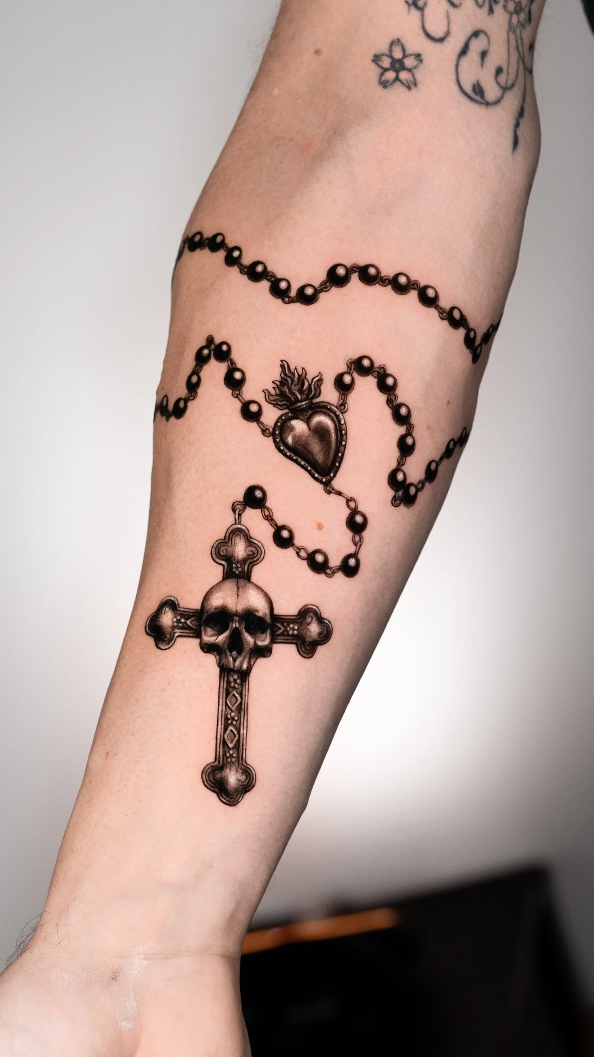 Tattooed forearm with a skull cross, rosary beads, and a heart with flame design.