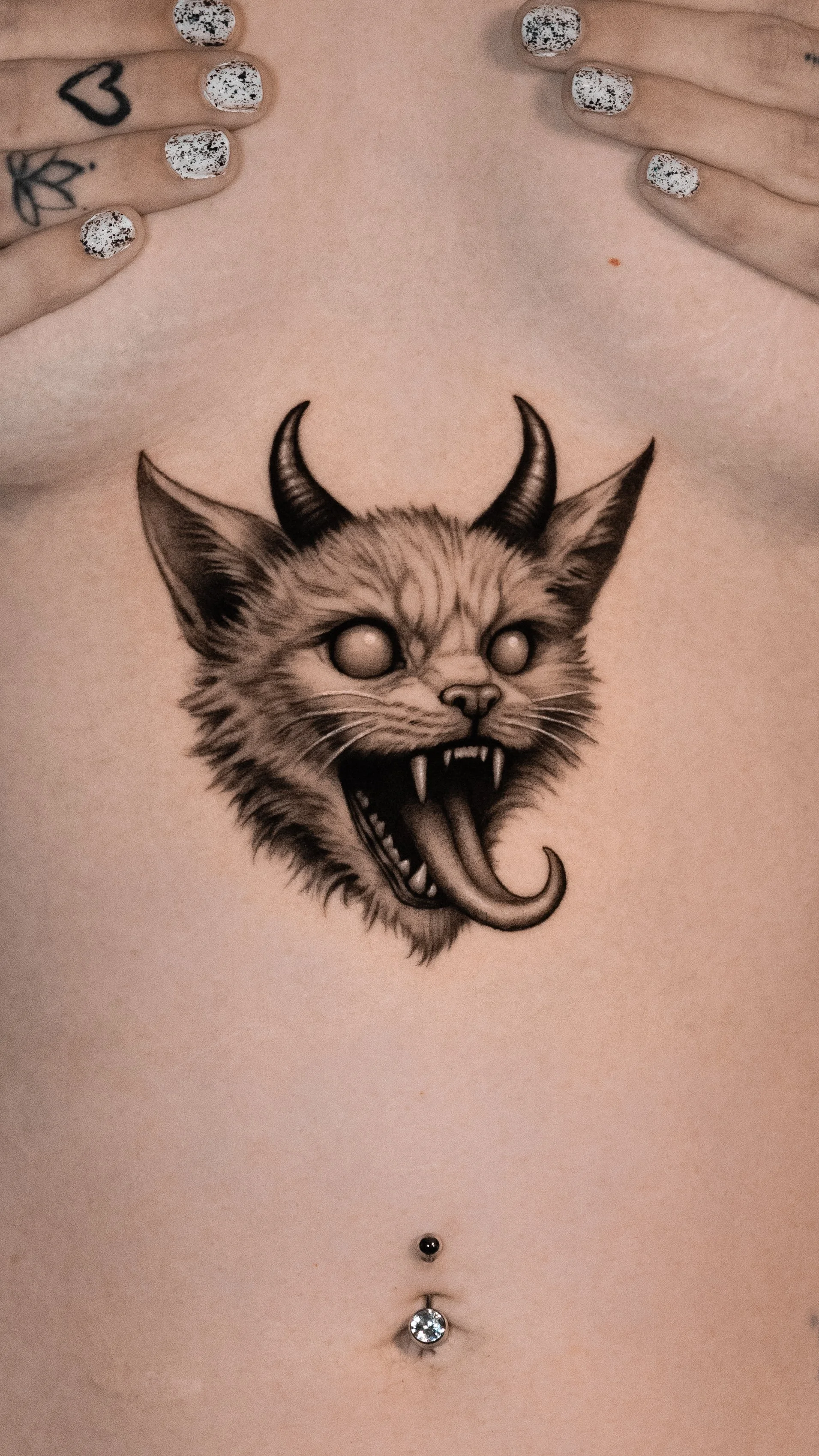 A tattoo of a snarling animal face with horns on a person's upper chest, with hands above the tattoo, showcasing heart and floral tattoos on fingers, and a pierced navel with a jeweled belly button piercing.