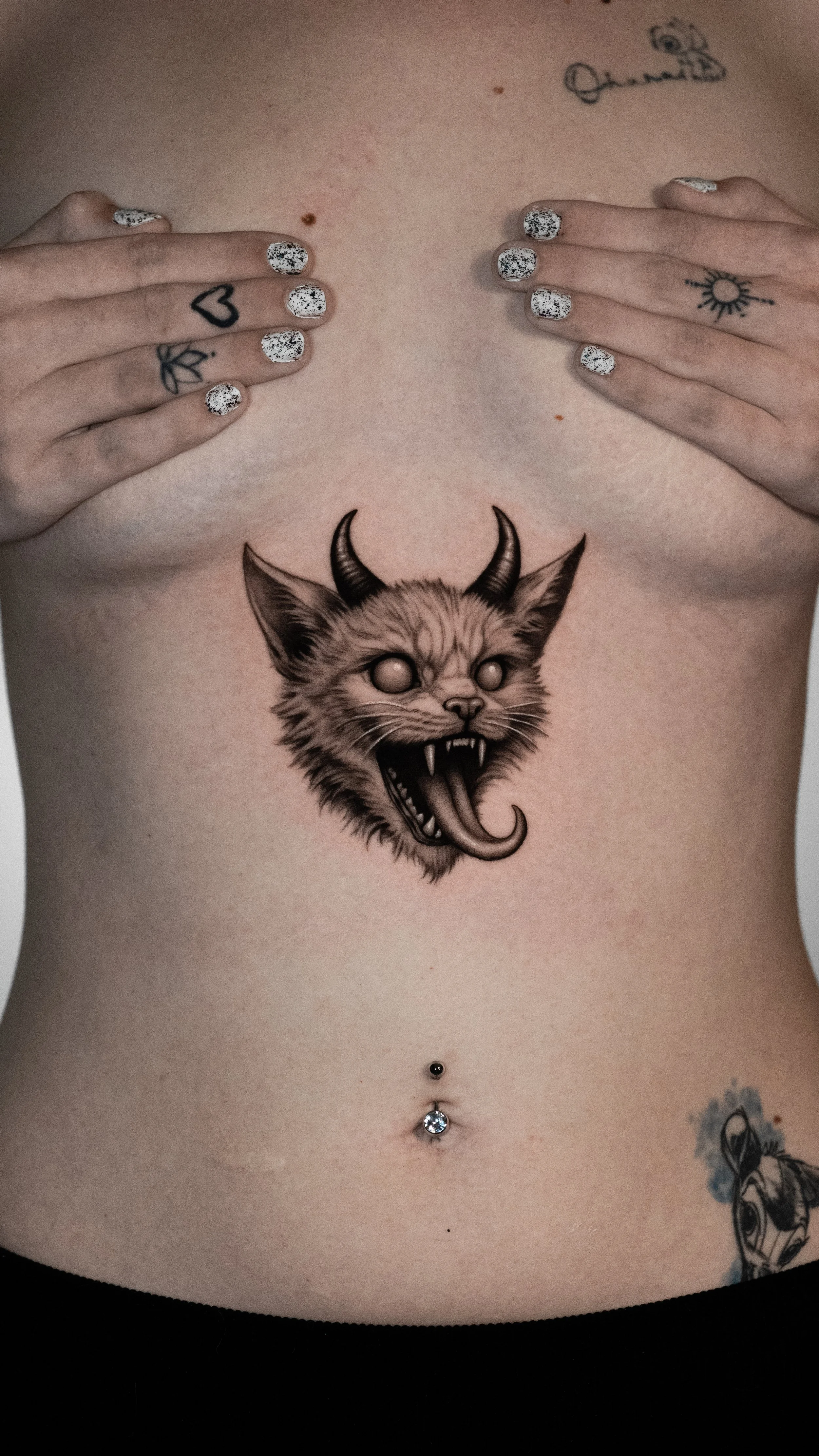 Close-up of a person's bare chest with a large black and grey tattoo of a snarling animal with horns and a long tongue. The person covers their breasts with their hands, which have speckled black and white nail polish and small tattoos of a heart and