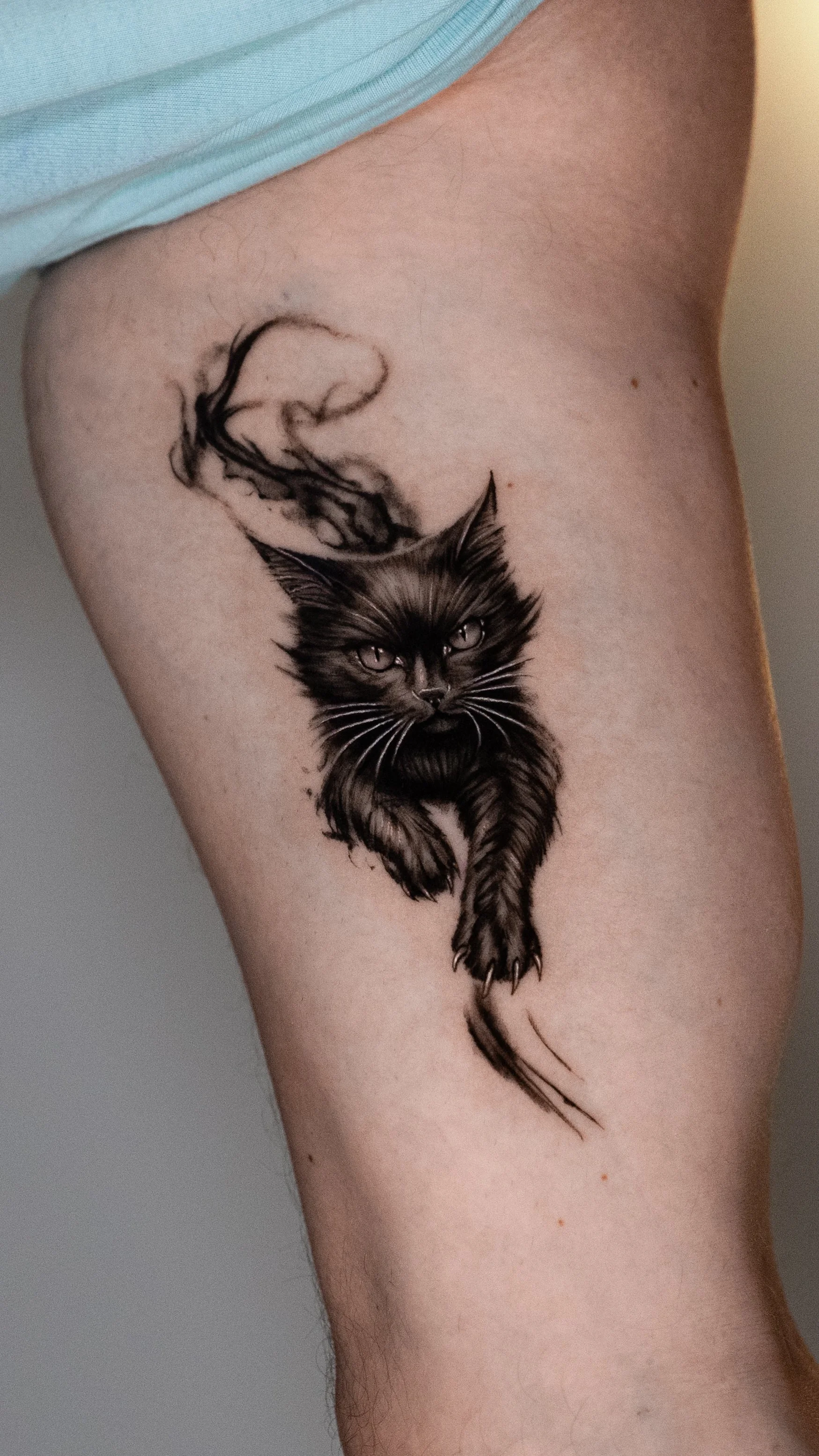 A tattoo of a black and gray cat with piercing eyes, sitting with its tail curling around its body, on a person's upper leg.