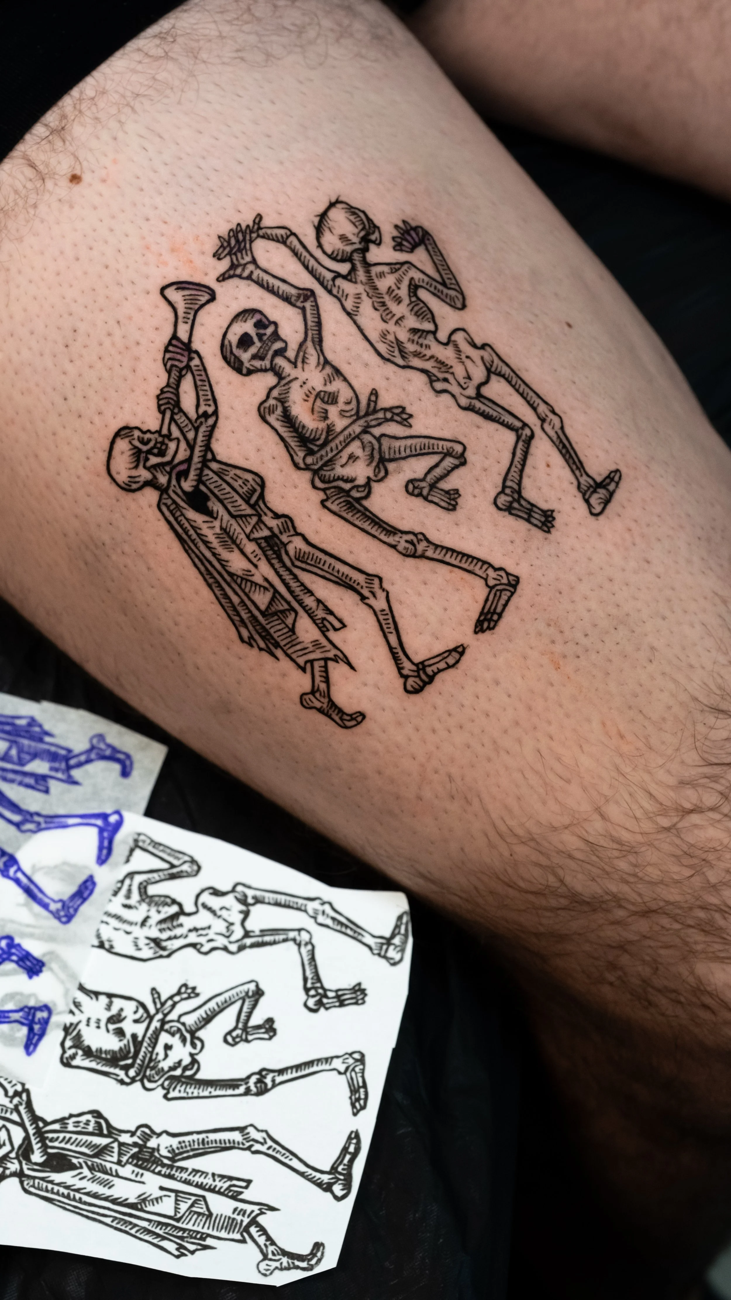 Tattoo of skeletons dancing holding hands on person's leg.
