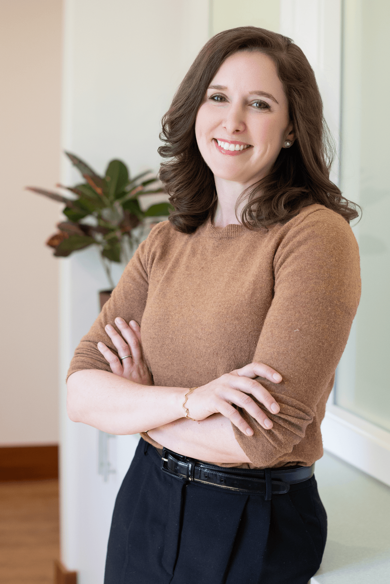 A smiling woman with shoulder-length brown hair, wearing a brown long-sleeved top and black pants, standing indoors with her arms crossed in front of her, near a potted plant and a window.