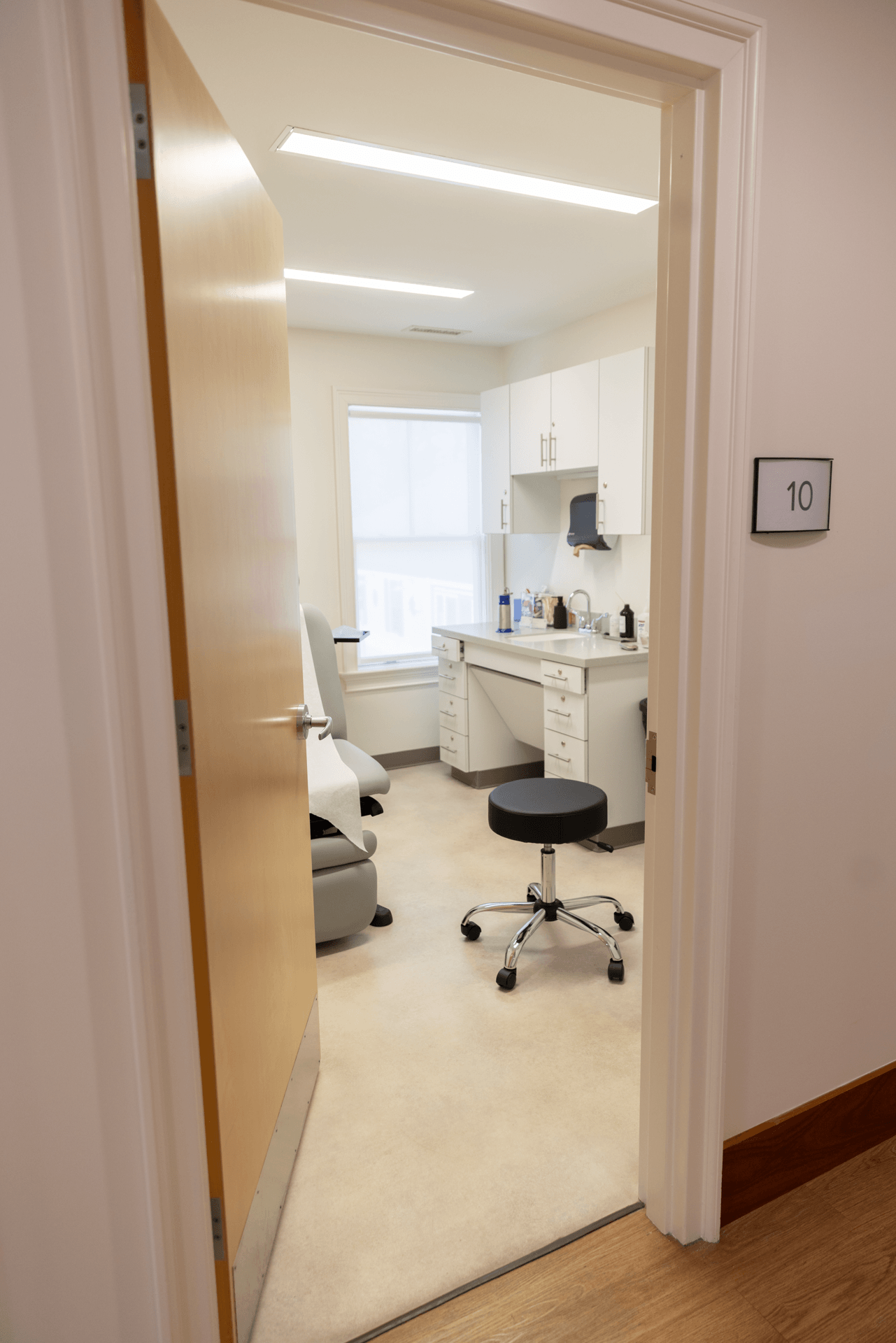 View through open door into a medical or examination room with a medical chair, desk, cabinets, and medical supplies, number 10 on the door.