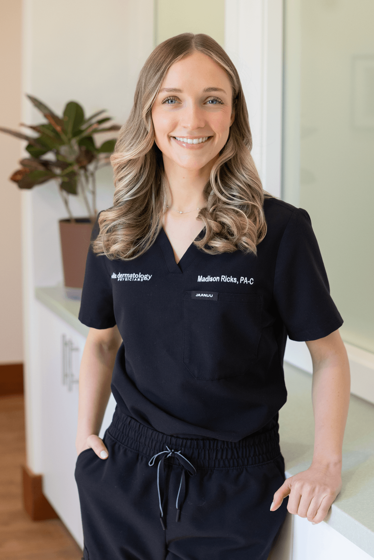 A woman in a navy medical uniform with embroidered name and organization, smiling and standing in a clinic or medical office setting with a plant in the background.