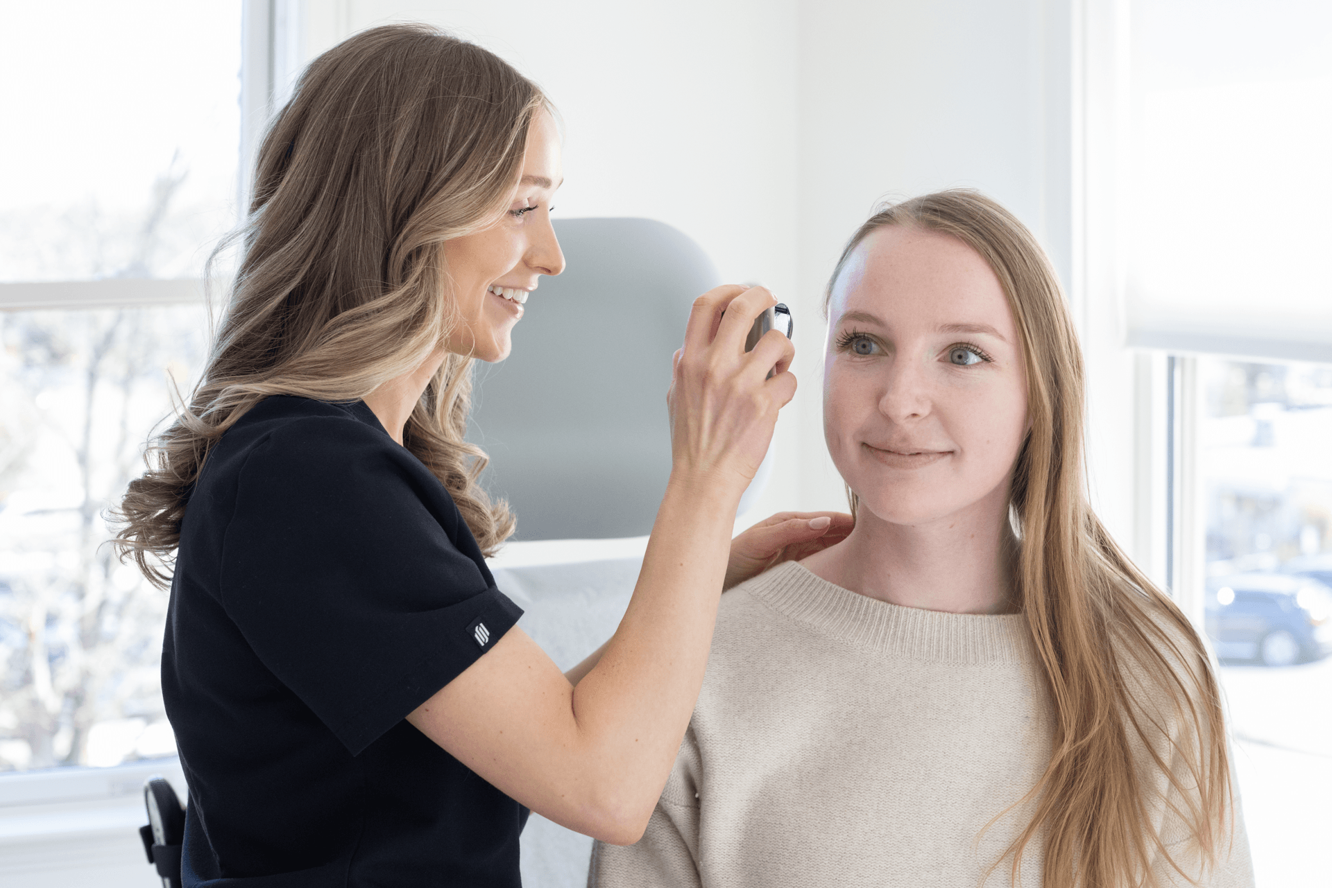 A woman with long blonde hair, dressed in a navy polo shirt, is smiling as she uses a small mirror to examine the face of a young woman with long red hair, wearing a beige sweater. They are in a bright room with large windows.