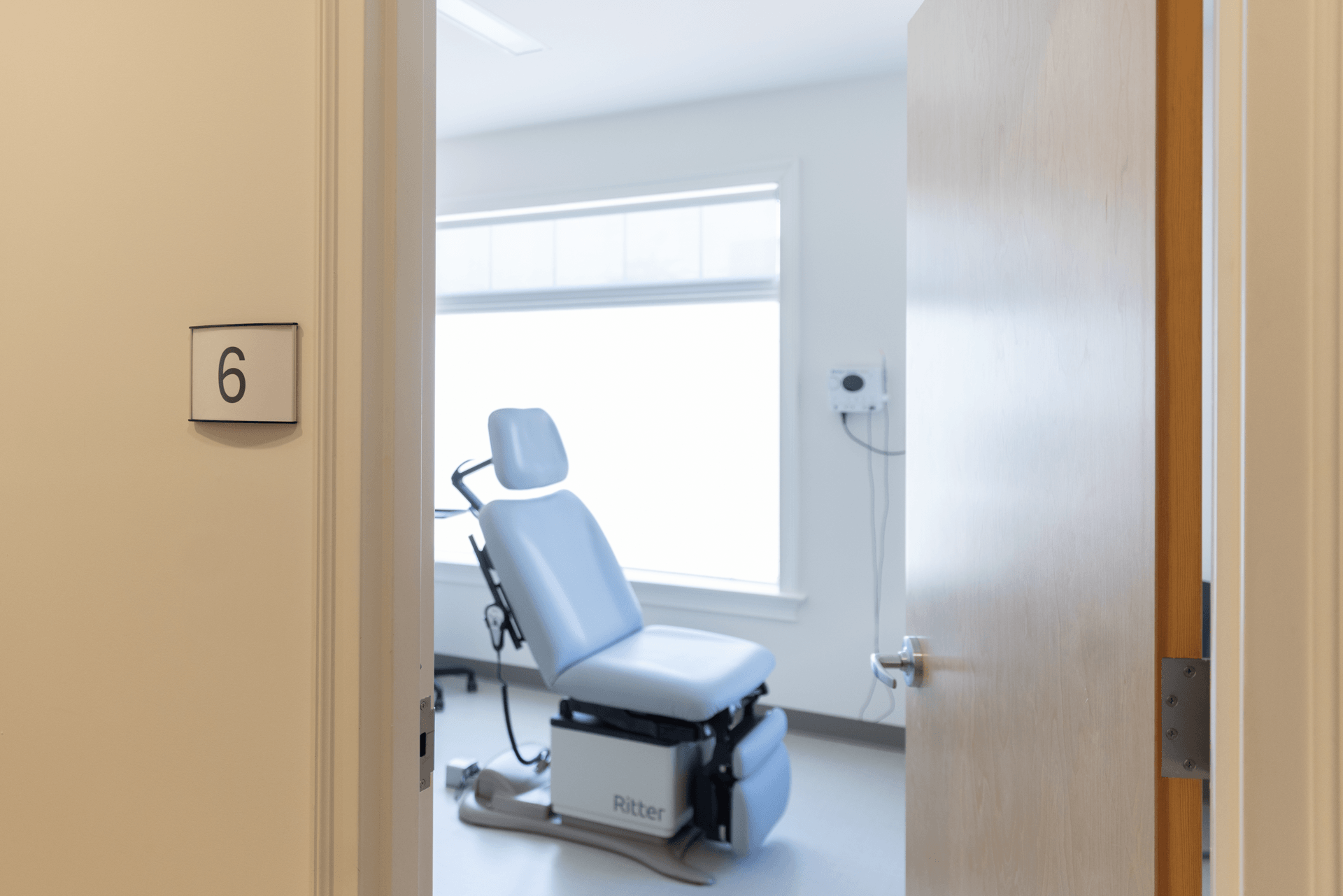 An empty dental examination room with a dental chair near a window, viewed through a partially open door labeled with the number 6.