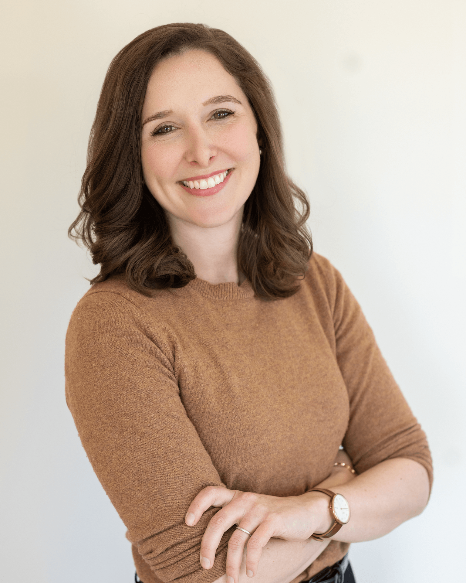A woman with brown hair in loose waves smiling with arms crossed, wearing a brown sweater and a watch.