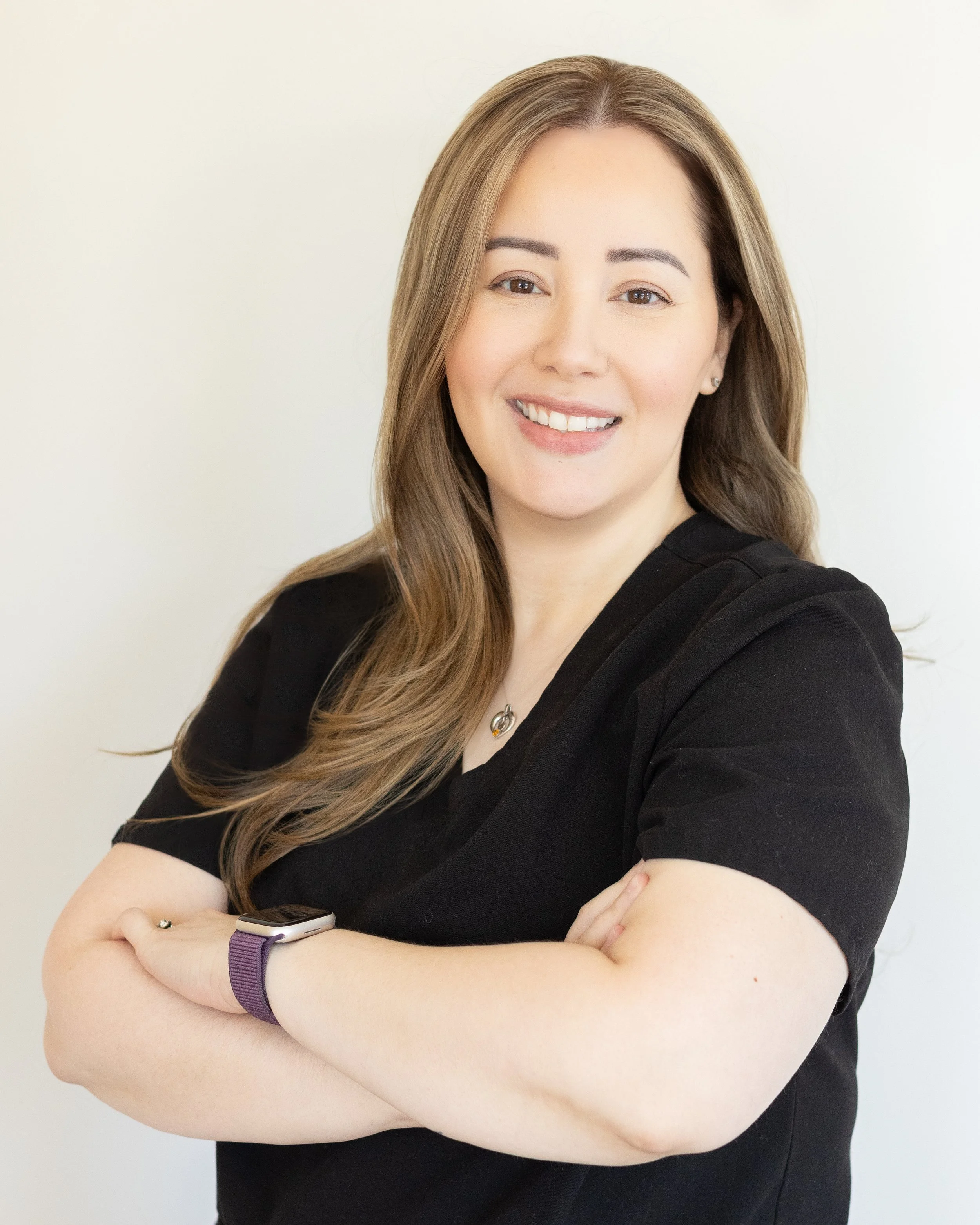 A smiling young woman with light brown hair, wearing a black top, gold earrings, and a purple smartwatch, with crossed arms against a plain white wall.