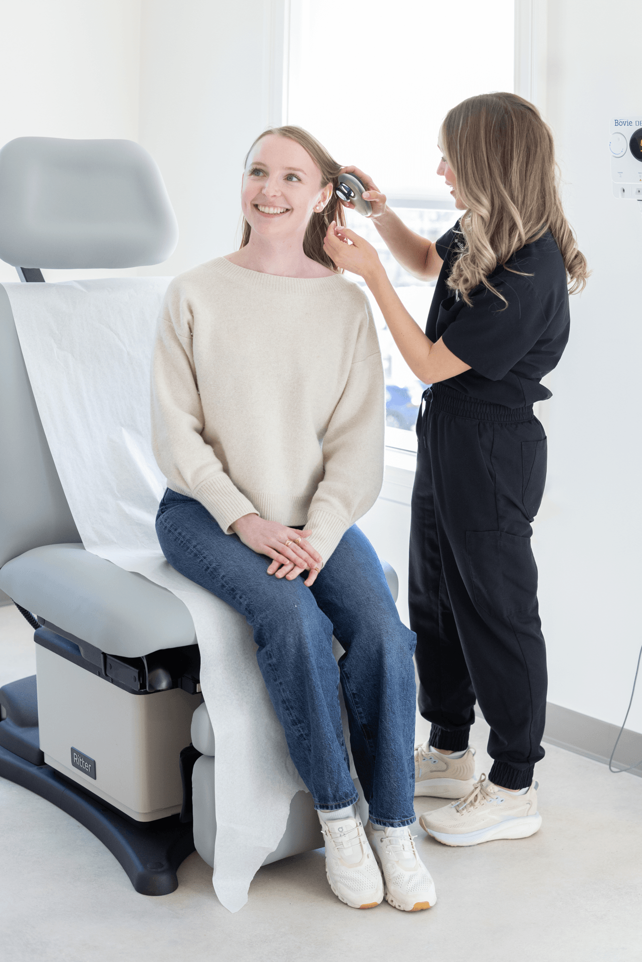 A dermatologist examines a woman in a white sweater sitting on a medical office chair smiling.