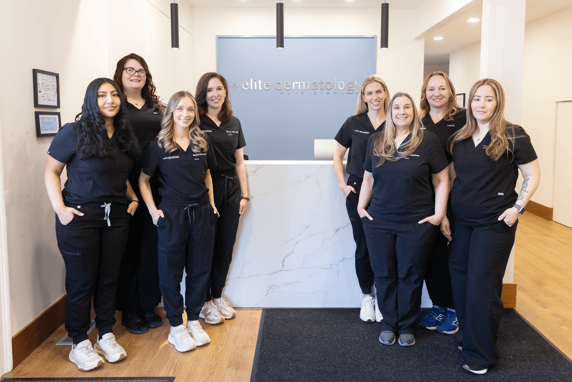 Group of nine women staff members standing in front of a reception desk at Elite Dermatology clinic, all wearing black scrubs and smiling.