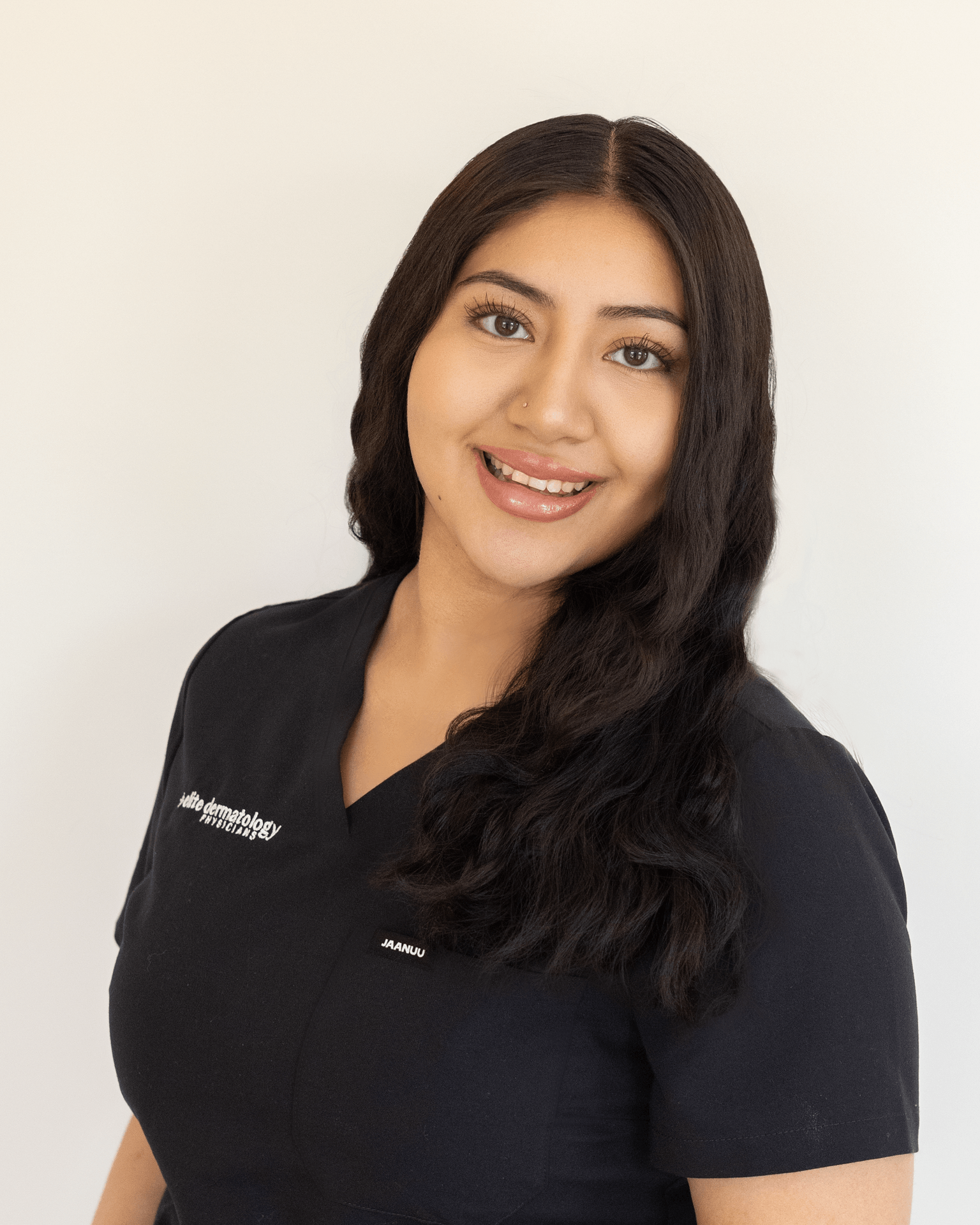 A woman with long dark hair and light skin smiling, wearing a black medical uniform with white text on it, standing against a plain light background.