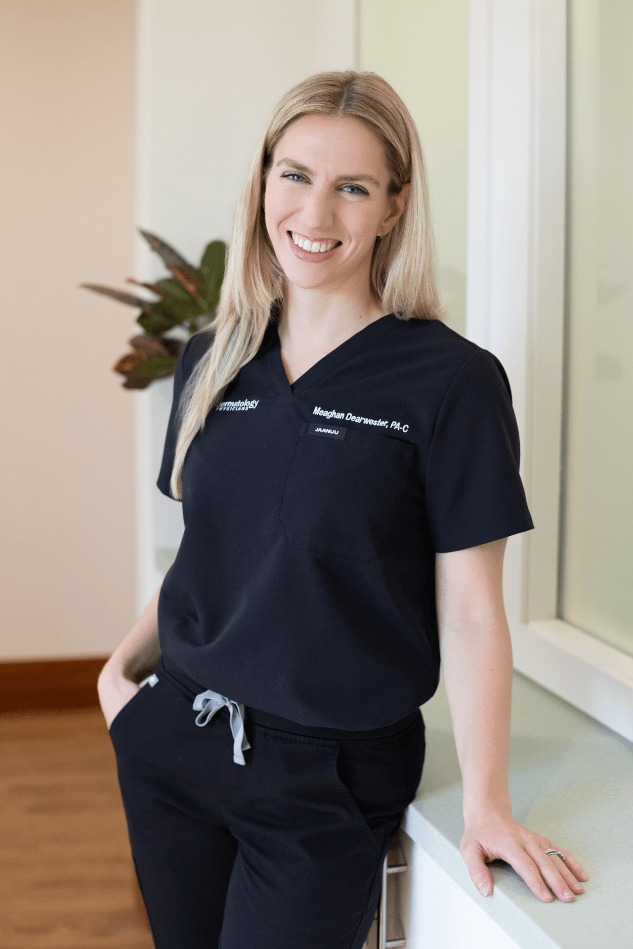 A woman with blonde hair wearing a navy scrub top with her name and title, smiling and standing with one hand resting on a white countertop in a room with light-colored walls and a potted plant in the background.
