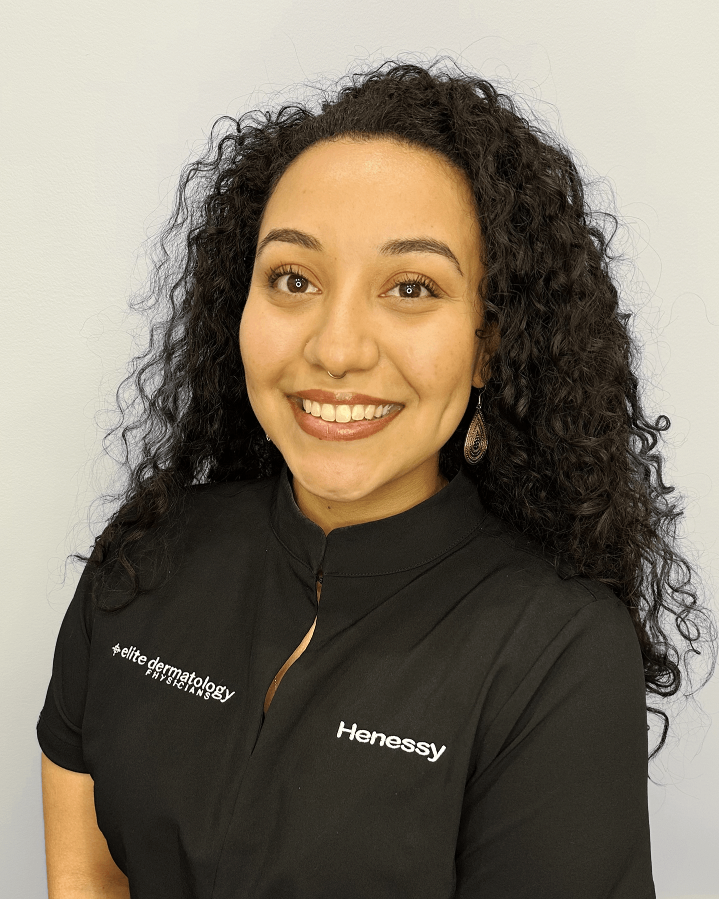 A woman with curly dark hair and light brown eyes smiling, wearing a black shirt with logo and text for Hennessy and elite dermatology, standing against a plain light-colored wall.