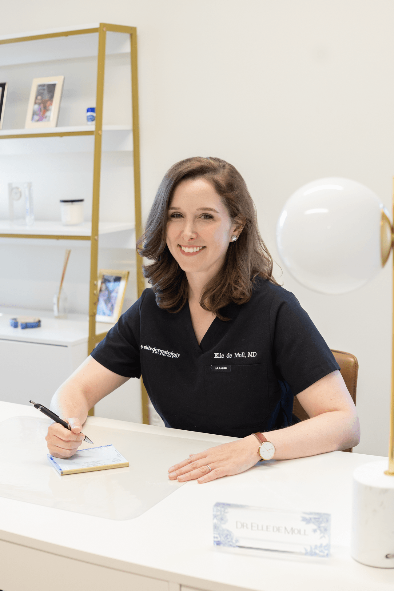 A woman with brown hair sitting at a white desk, smiling, wearing a black medical uniform with a badge reading Elle de Moll, MD, in a room with a white wall and shelves with framed photos and small objects.