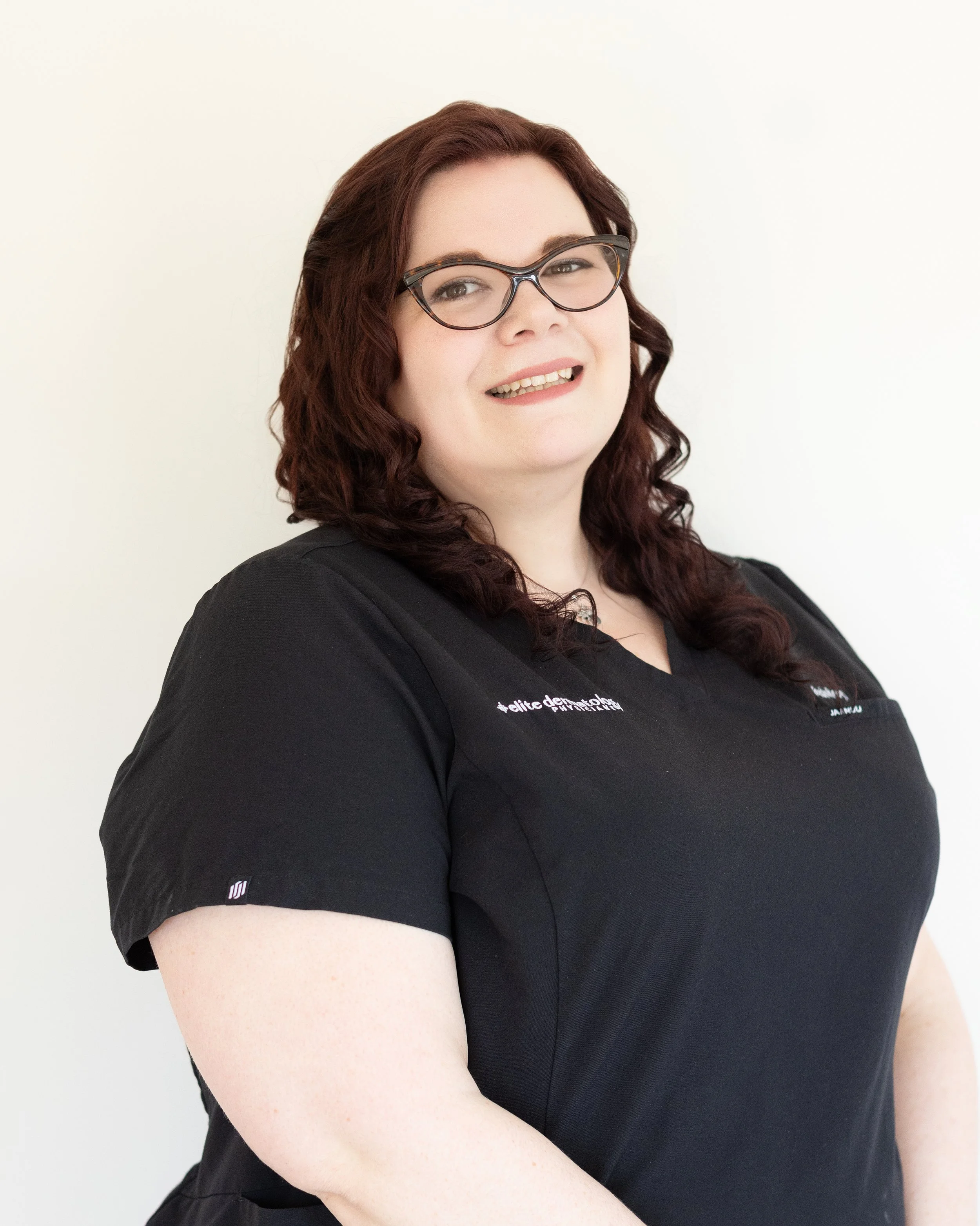 Portrait of a woman with brown curly hair, glasses, smiling, wearing a black shirt, against a plain light background.